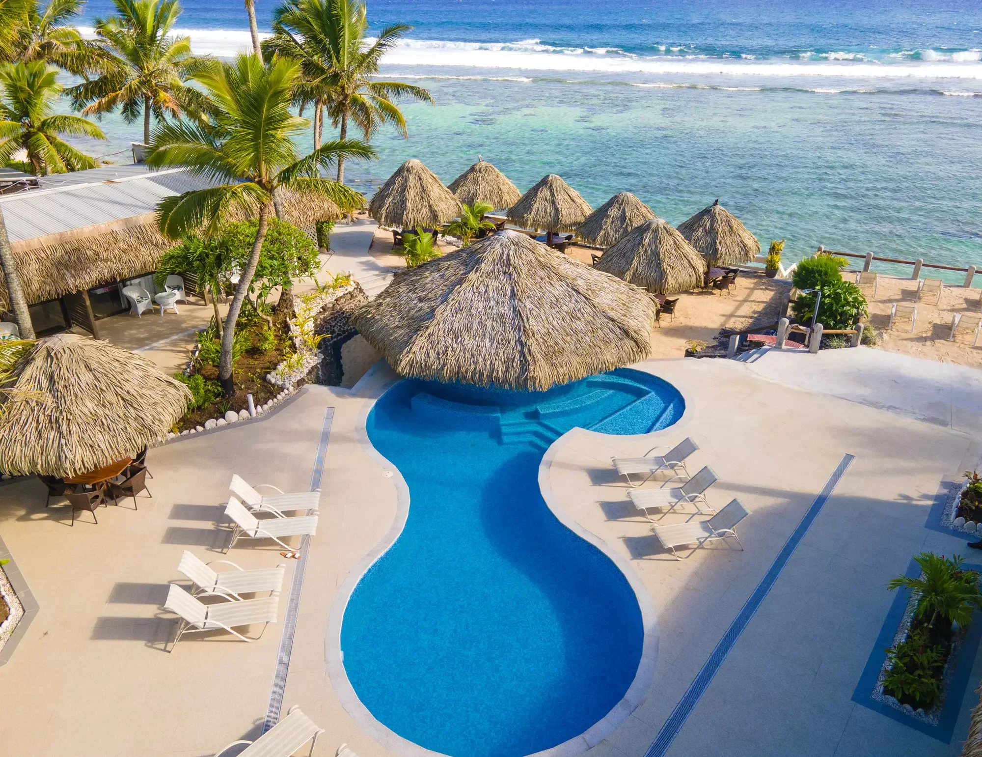 A tropical beachfront resort with a swimming pool, thatched-roof huts, lounge chairs, palm trees, and access to the ocean with waves.