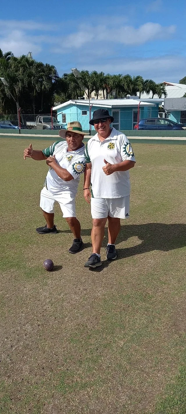 Two people on a lawn bowling green, both wearing white sports outfits with green and yellow accents, one giving a thumbs-up, and the other gesturing with a pointing finger. The scene is outdoors with a blue building, palm trees, and parked cars in the background.