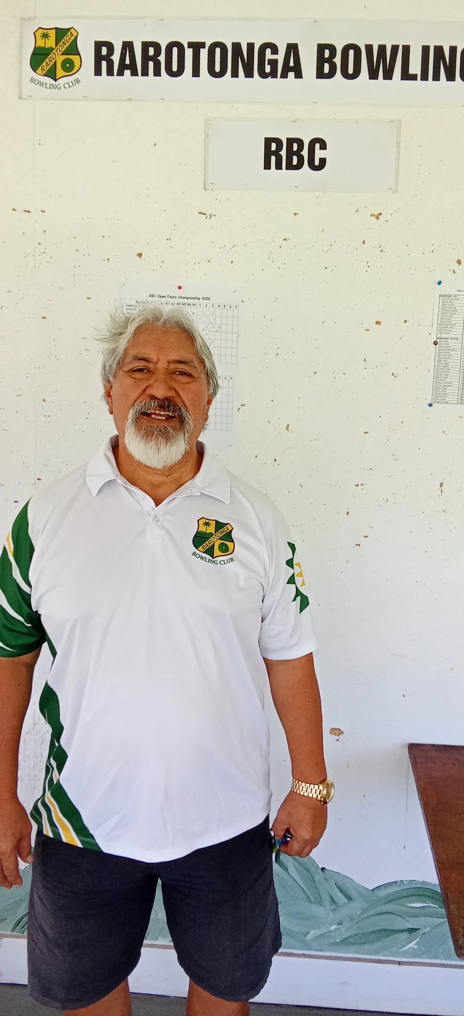 A man wearing a white Rarotonga Bowling Club polo shirt and black shorts standing indoors in front of a white wall with signs that say 'Rarotonga Bowling Club' and 'RBC.'