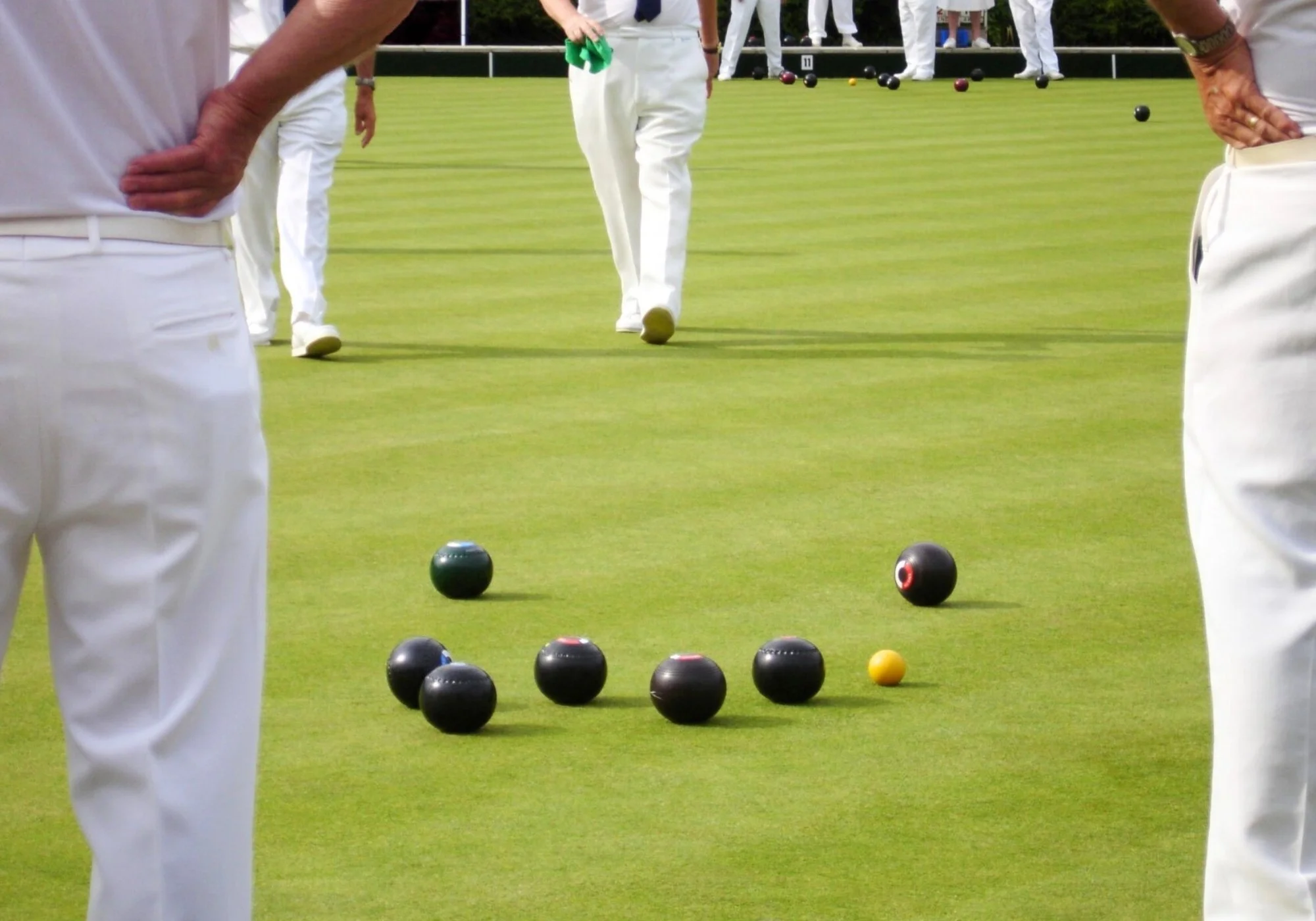 Lawn bowls game on a green field with players in white clothing, black bowls, a yellow jack, and people standing in the background.