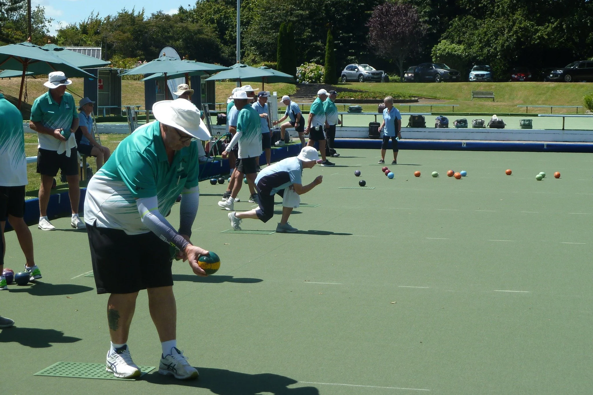 A group of seniors playing lawn bowls outdoors on a sunny day, with some preparing to bowl and others observing under umbrellas.