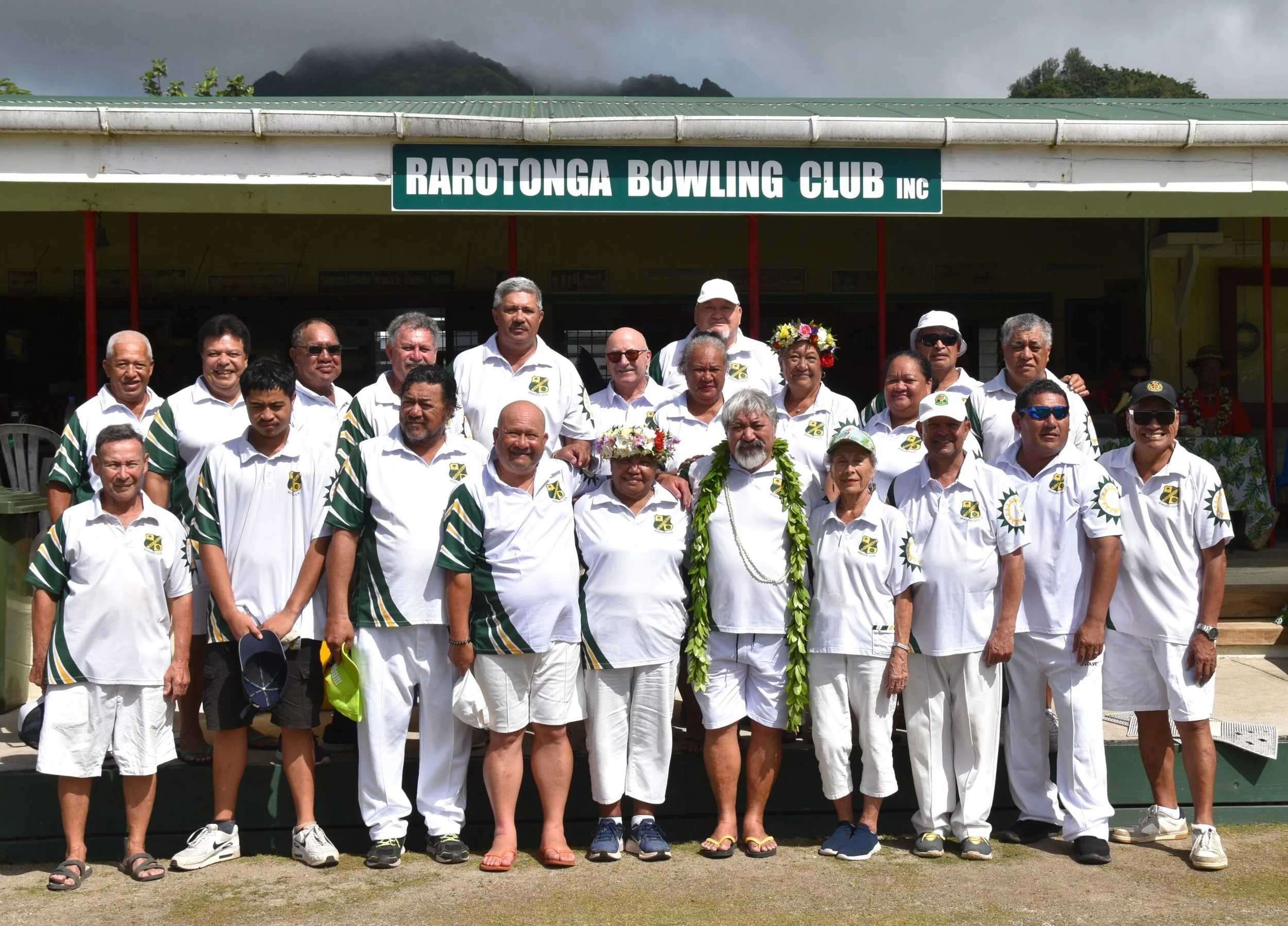 Group of people posing in front of Rarotonga Bowling Club building, some wearing floral crowns and leis, with mountains in the background.
