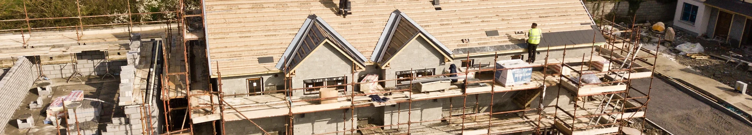 Construction site with workers building a house, scaffolding around the structure, and a roof with sections being installed.