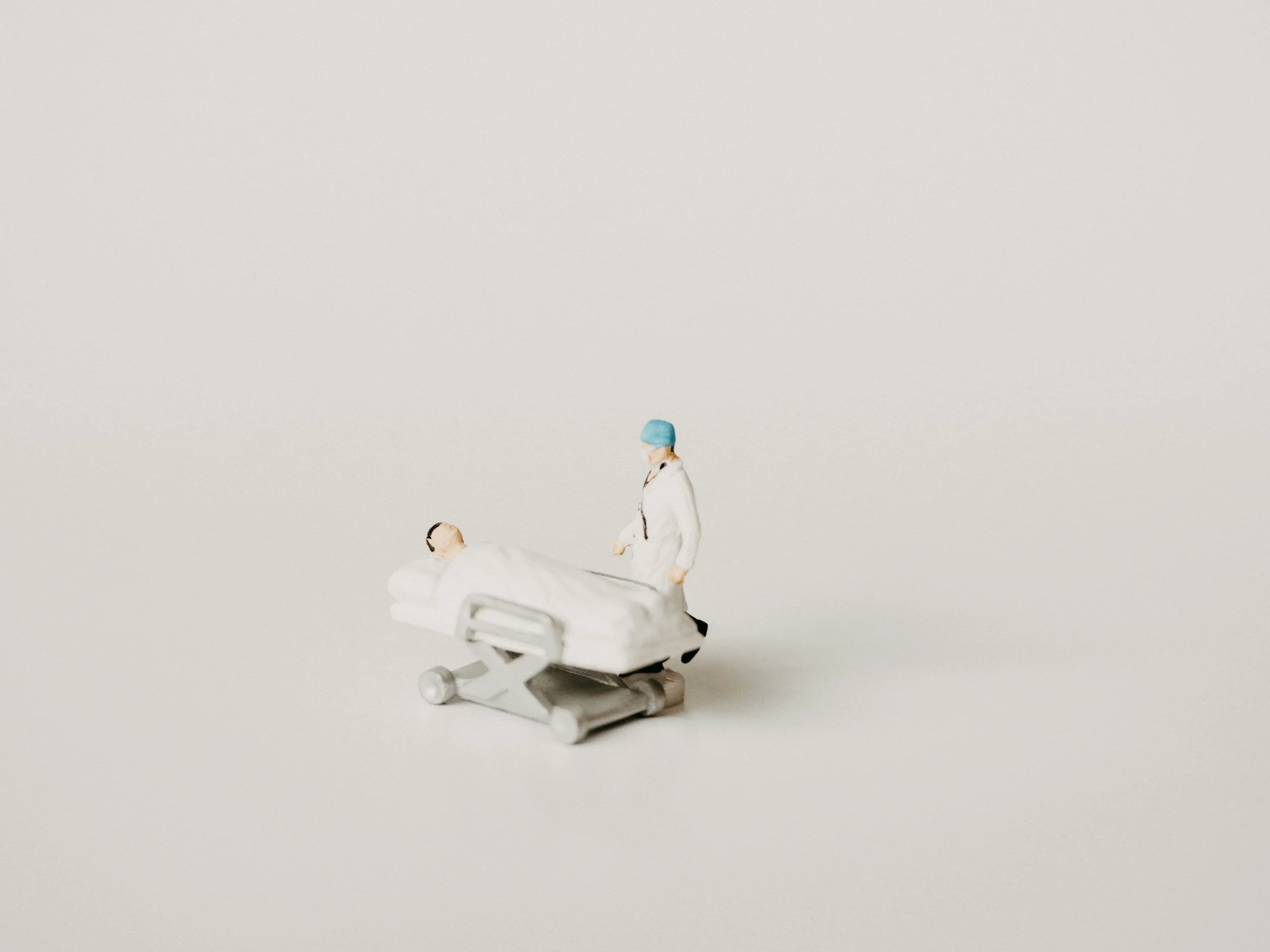 Miniature doctor figurine attending to a patient in a hospital bed on a plain white background.