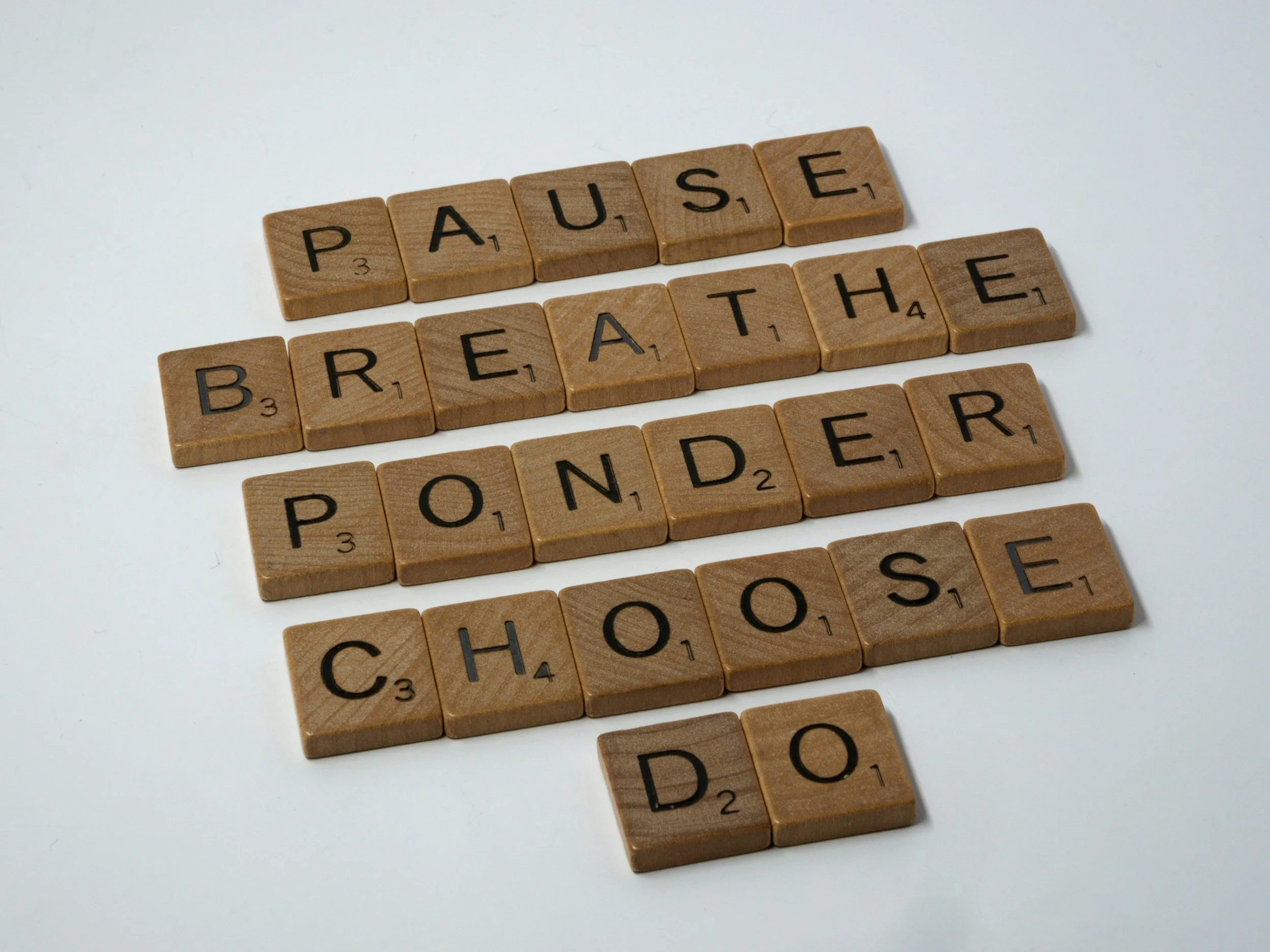 Wooden Scrabble tiles spelling out "PAUSE," "BREATHE," "PONDER," "CHOOSE," and "DO" on a white background.