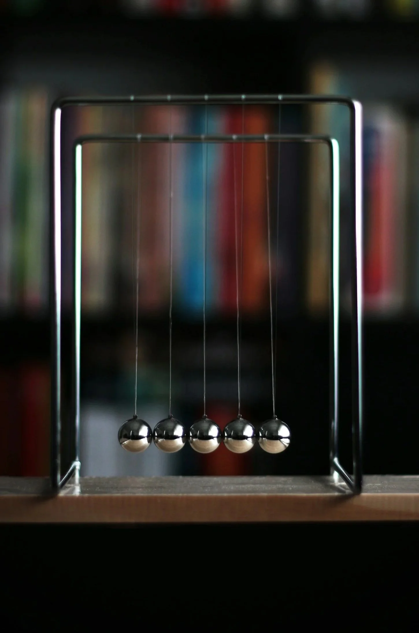 Newton's cradle with five metal balls swinging in motion on a wooden surface, with a blurred bookshelf background.