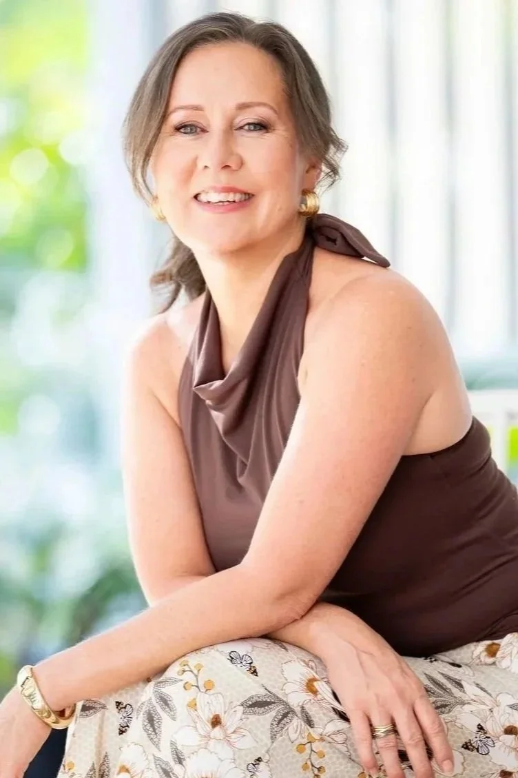 A smiling woman with short brown hair, wearing gold earrings, a brown sleeveless top, and a floral skirt, sitting with her arms crossed on a table indoors with a blurred green and white background.