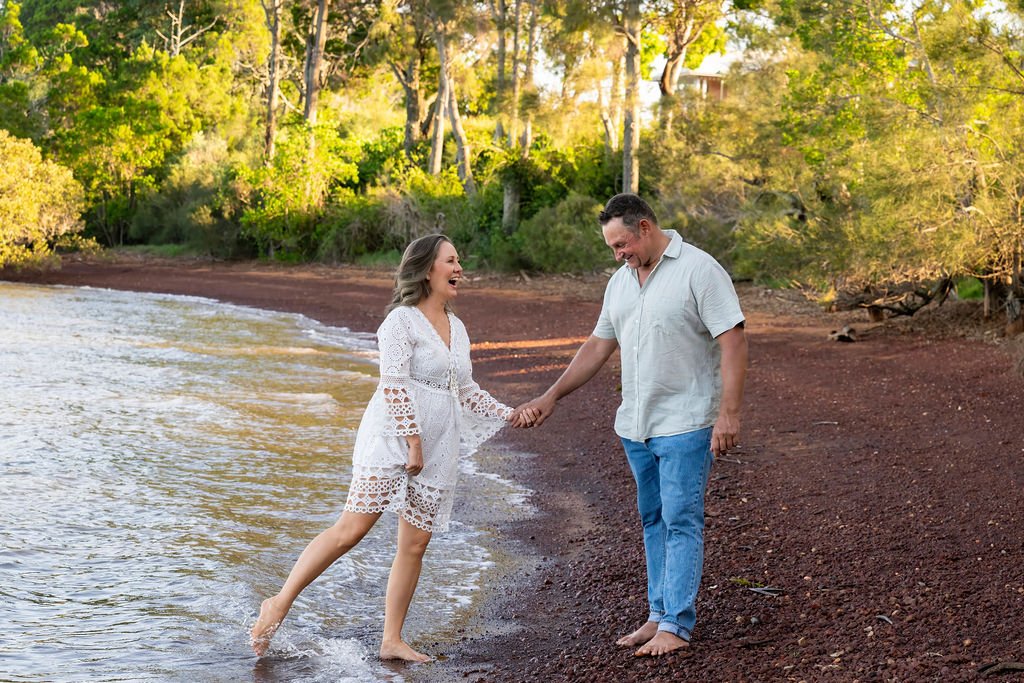 A couple holding hands and smiling on a beach with trees and sunlight in the background.