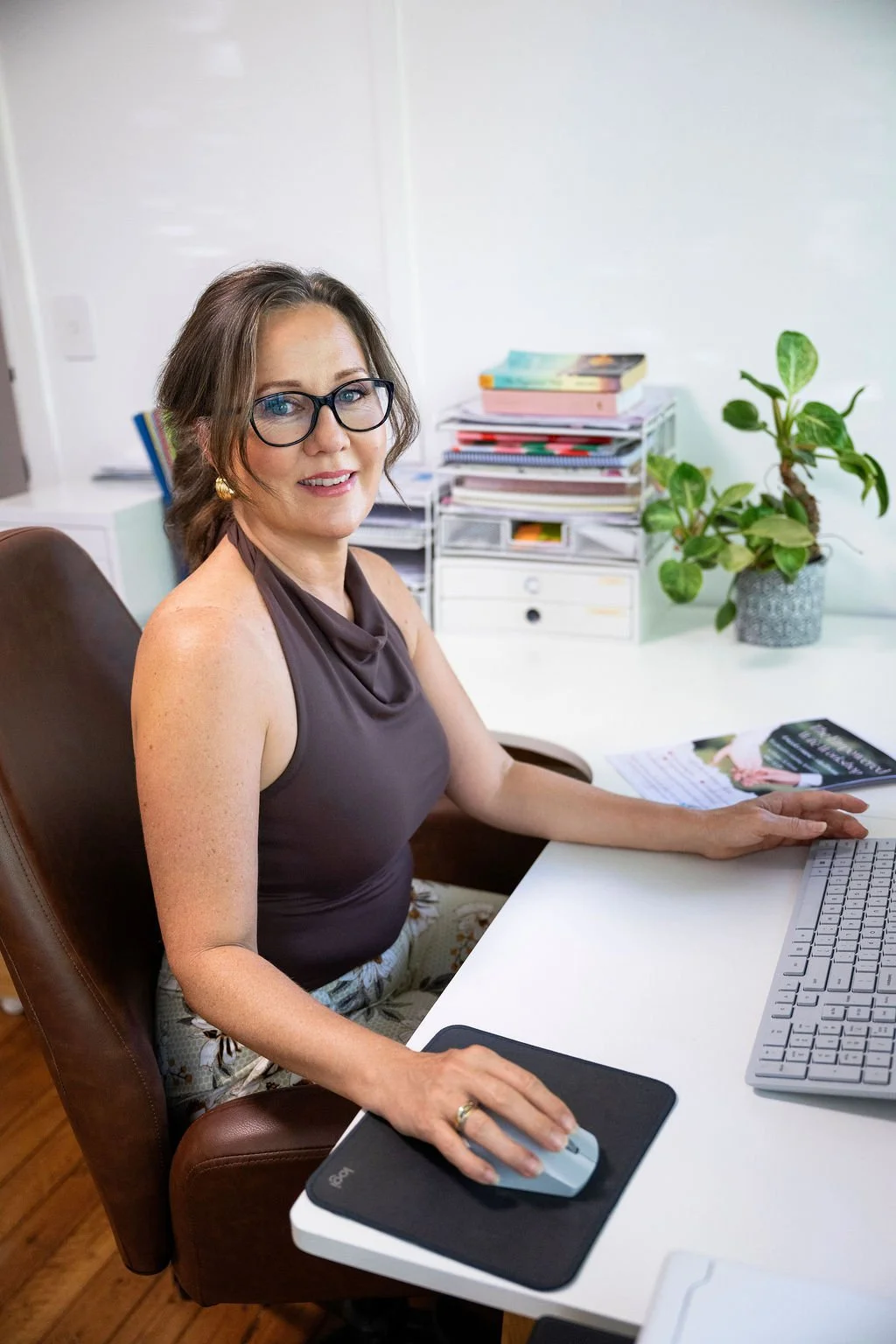 A woman with glasses and earrings sitting at a white desk in an office, working on a computer with a mouse on a mouse pad, with a stack of papers and books and a potted plant in the background.