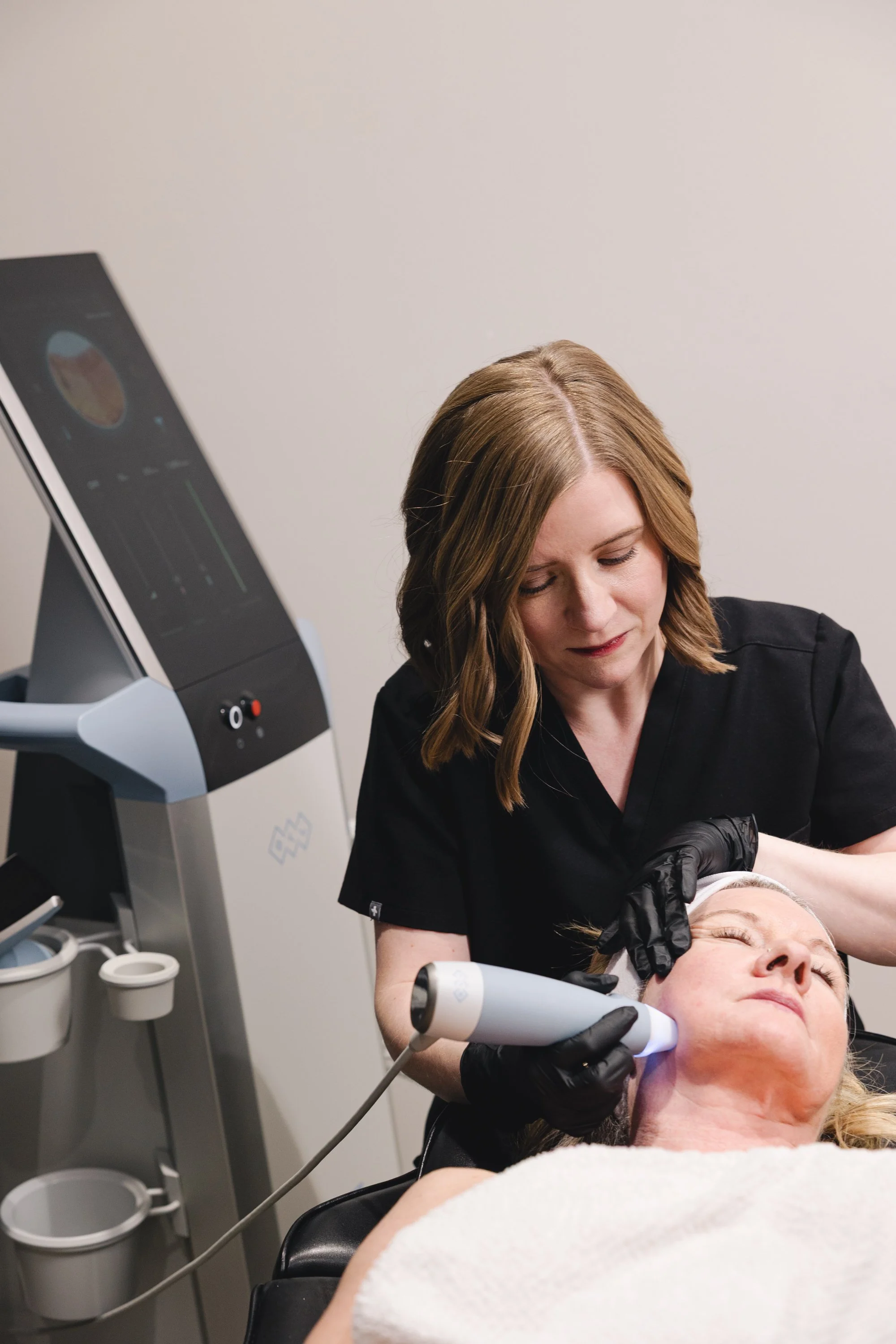 A woman with blonde hair receiving a facial treatment from a skincare professional wearing black gloves, in a clinical setting with advanced equipment nearby.