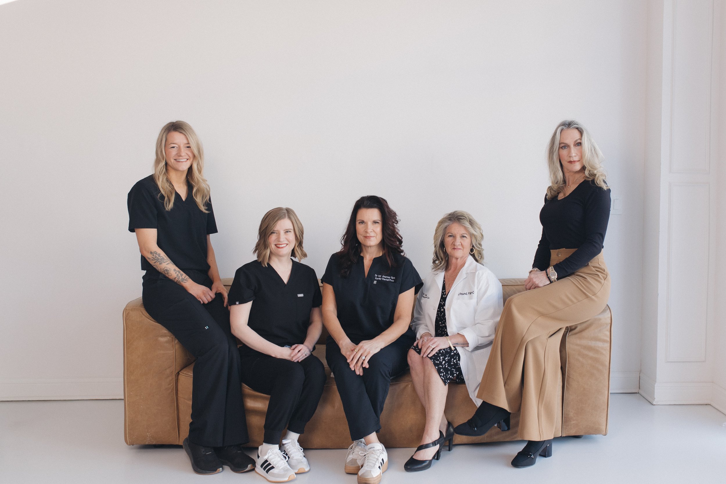 Group of six women, some wearing medical scrubs and one in a white coat, posing together inside a room with plain white walls, and a brown cushioned bench.