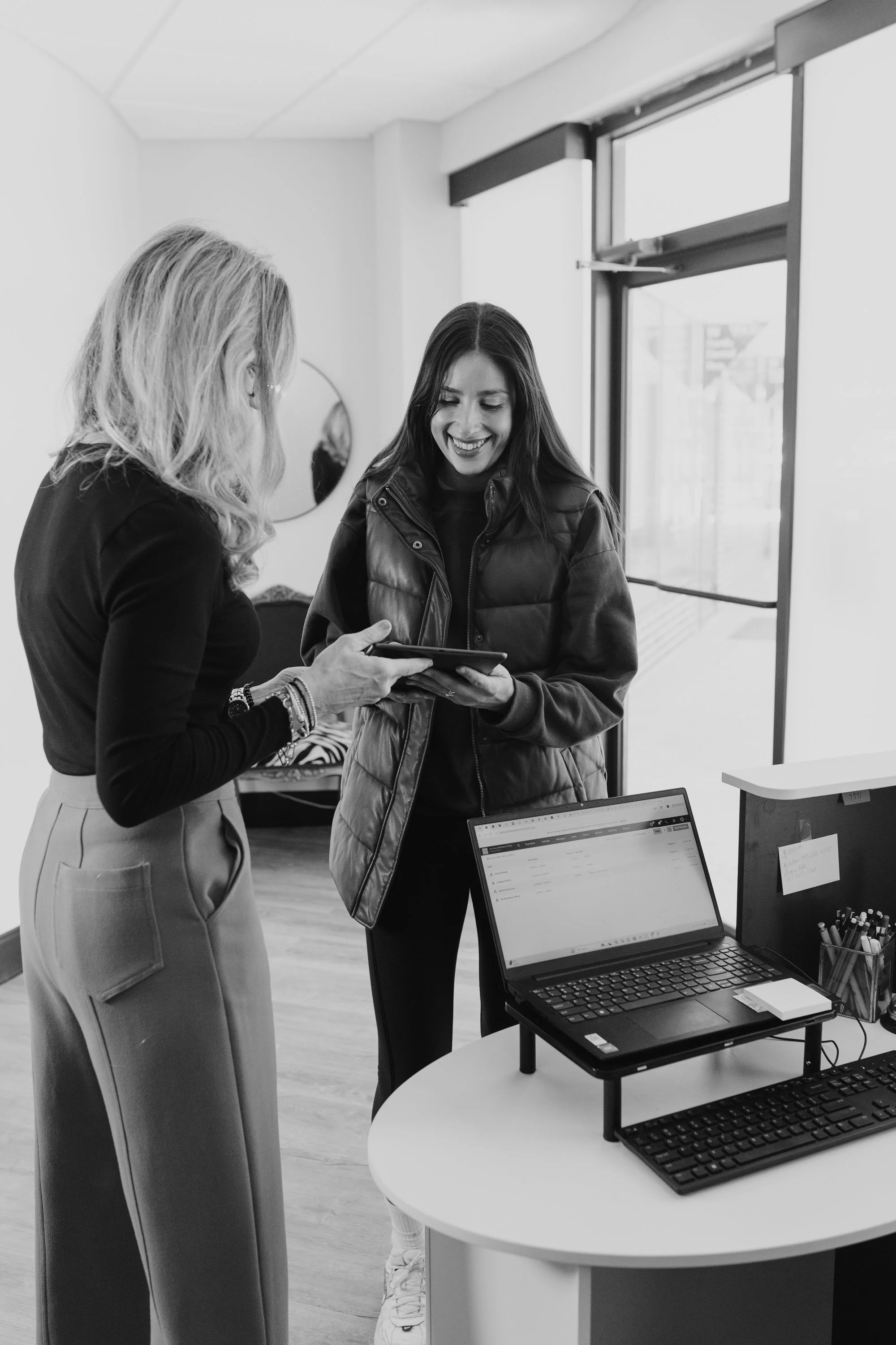 Two women standing by a desk with a laptop, keyboard, and printer, looking at a tablet in an office.