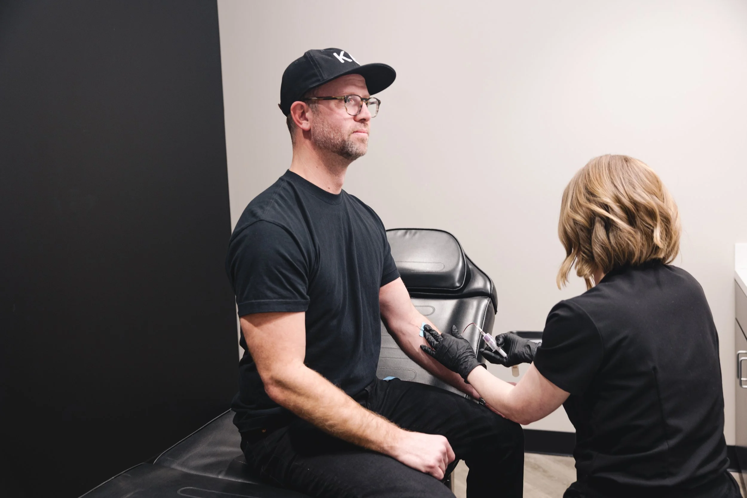 A man sitting on a black chair during a medical vaccination, with a woman in black scrubs administering a shot into his upper arm.