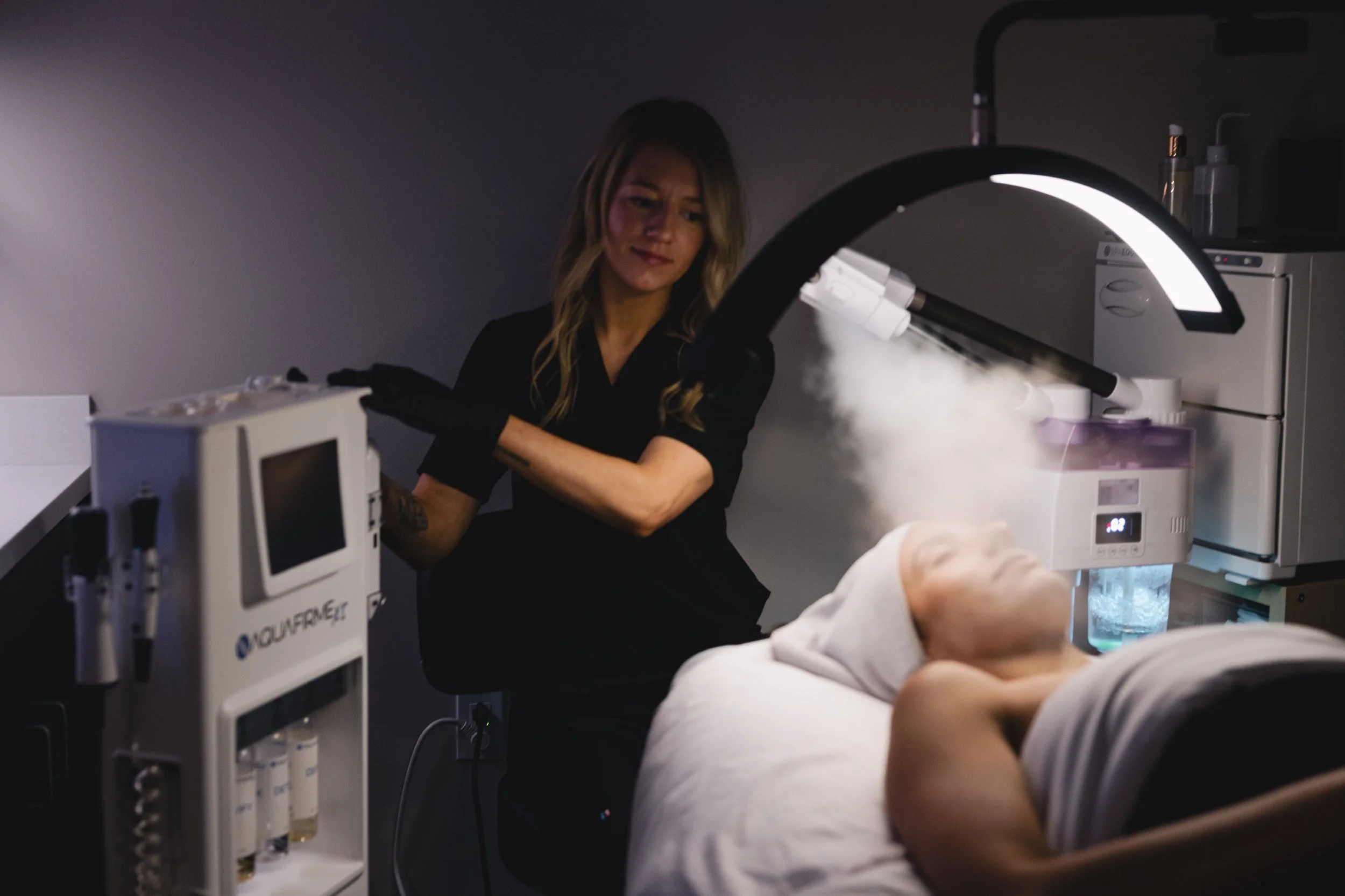 A woman using a medical or laboratory microscope in a dimly lit room, with medical equipment and a patient's arm visible in the foreground.