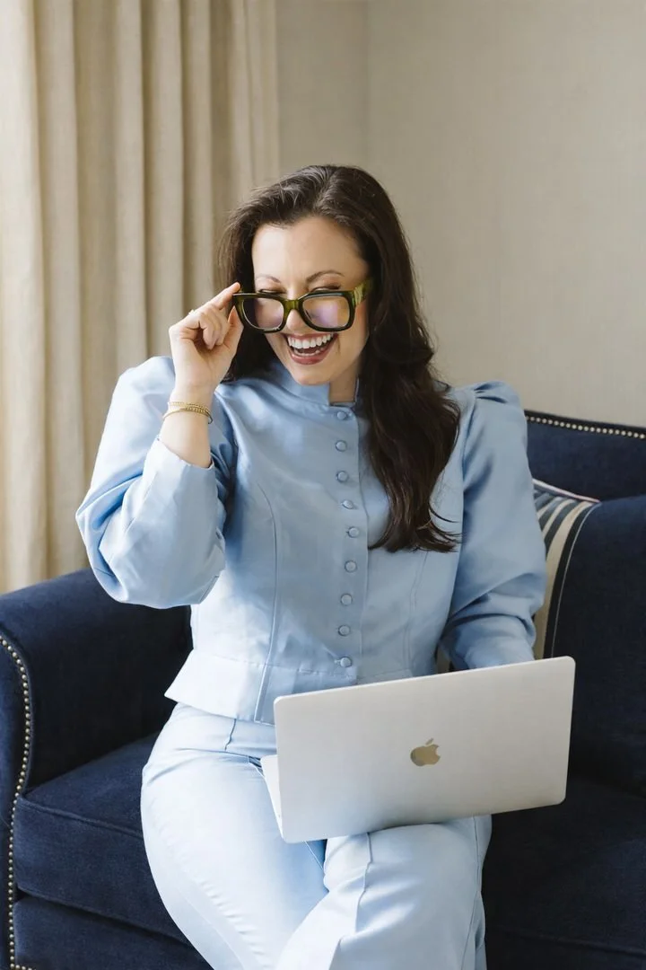 A woman with long dark hair, wearing glasses and a light blue outfit, smiling and looking at her laptop while sitting on a dark blue couch in a cozy room.