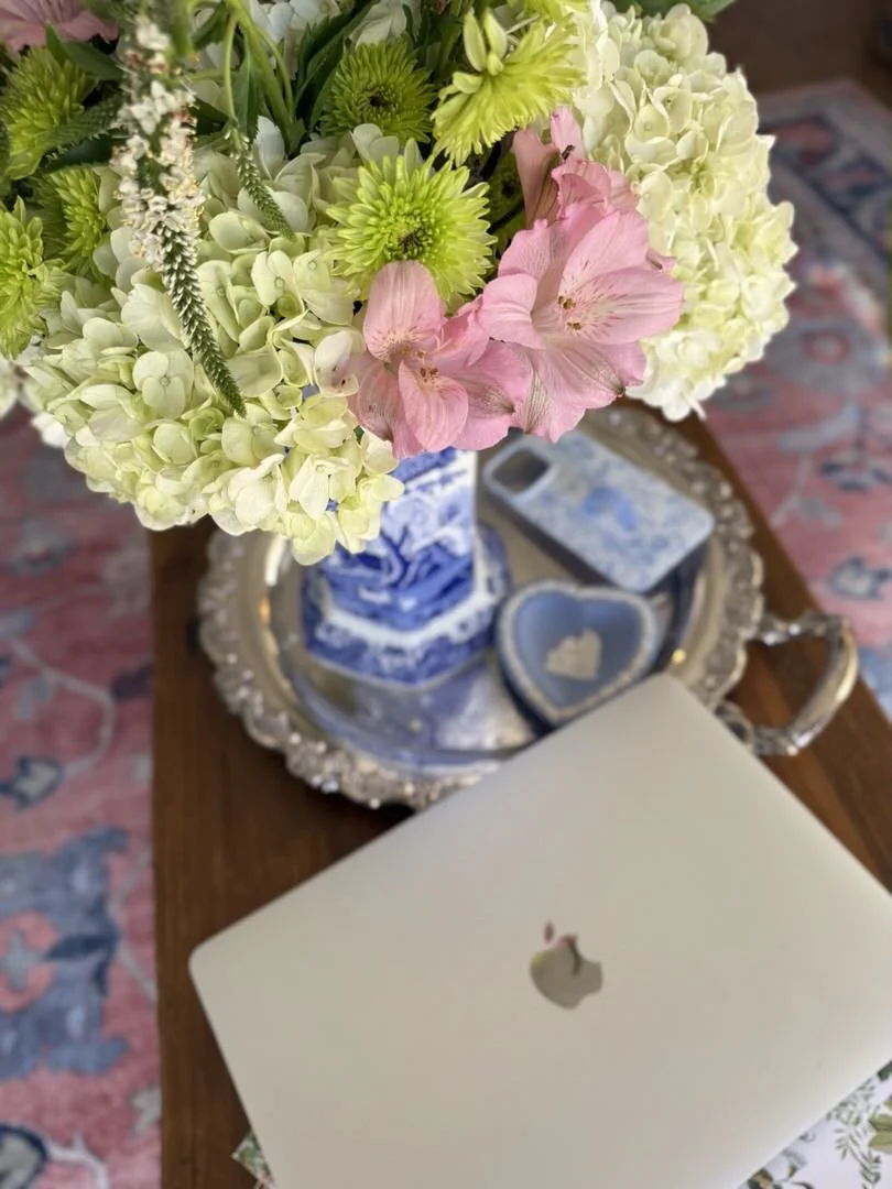 A bouquet of white hydrangeas, pink lilies, and green flowers on a silver tray next to a closed Apple MacBook and a decorative heart-shaped dish, on a wooden table.