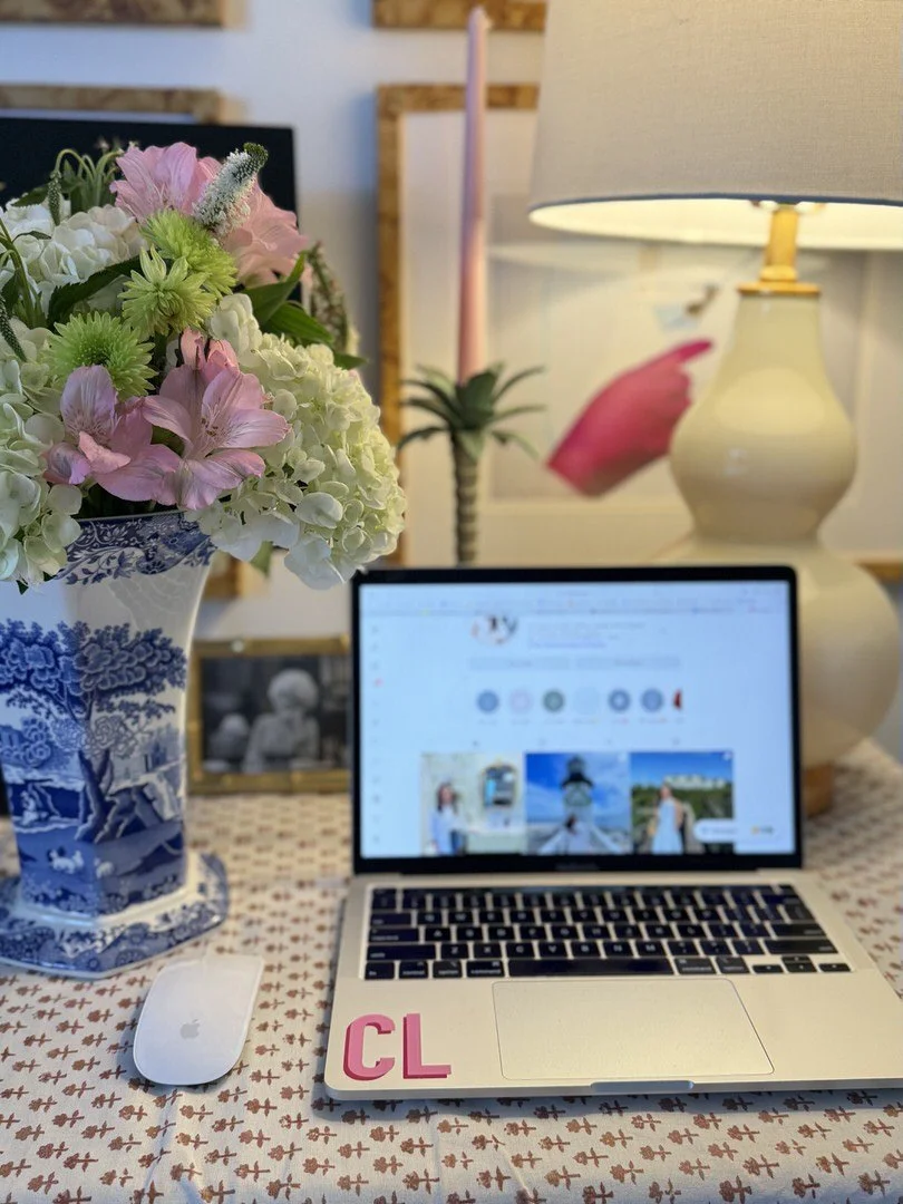 A desk with a vase of pink and white flowers, a MacBook laptop displaying a Pinterest page, a white Apple mouse, and a large beige table lamp, with framed artwork and decorative items in the background.