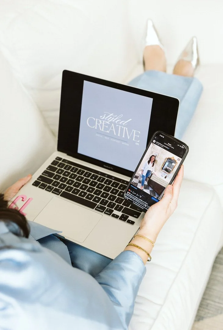 A woman sitting on a white couch, holding a smartphone showing her social media feed, with a laptop open on her lap displaying a graphic that says 'Styled Creative.' She is wearing a light blue satin shirt, white shoes, and gold bracelets.