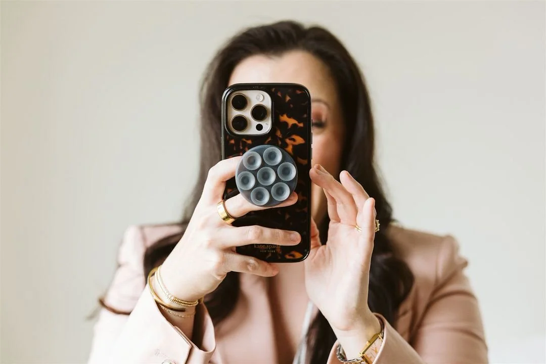 A woman with dark hair taking a selfie with a phone that has a pop socket attached, wearing a beige blazer and jewelry.