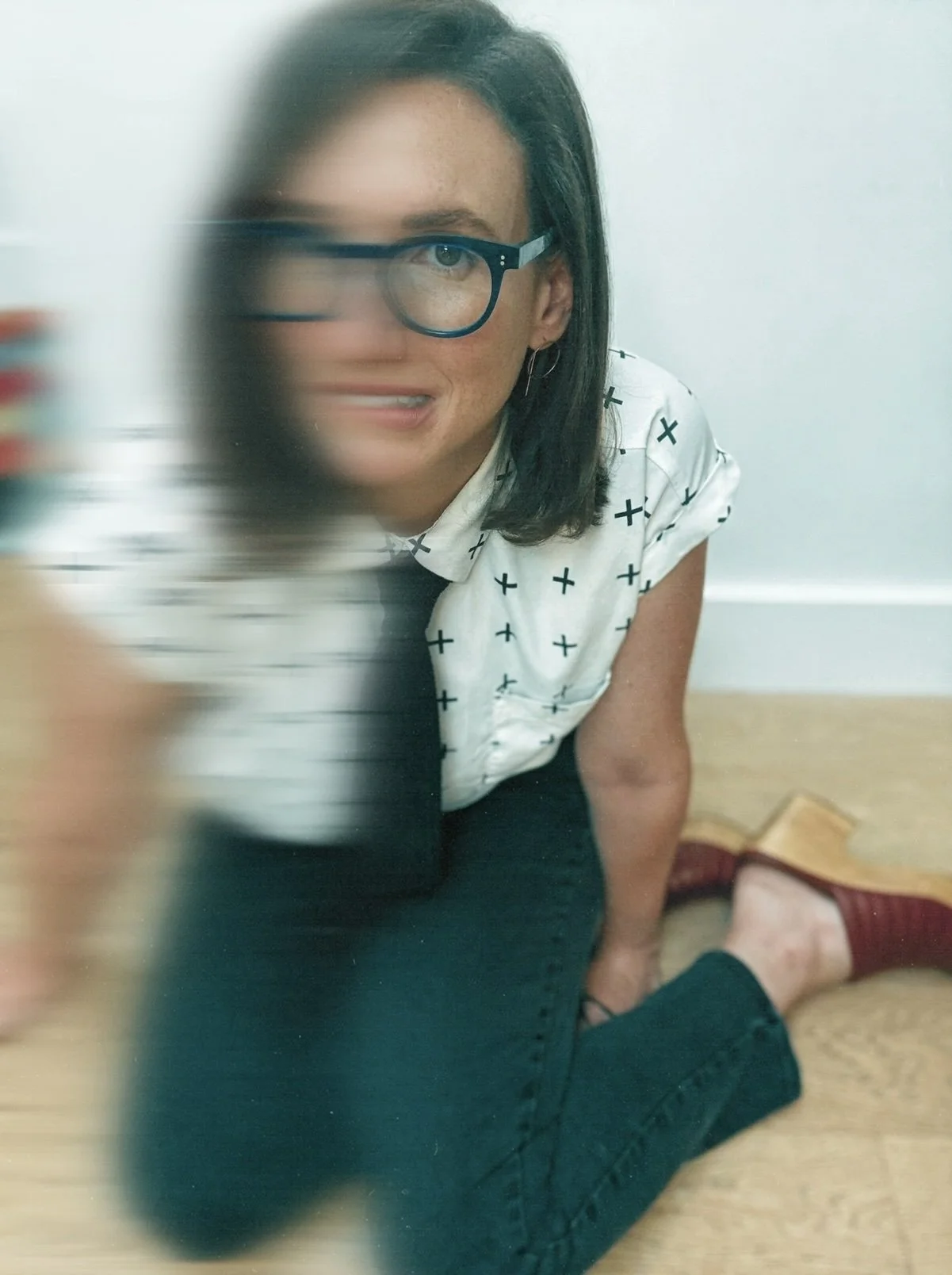 A woman with glasses and dark hair is sitting on the floor next to a white wall, wearing a white shirt with black crosses, black pants, and red shoes. The photo has a blurred effect on the left side.