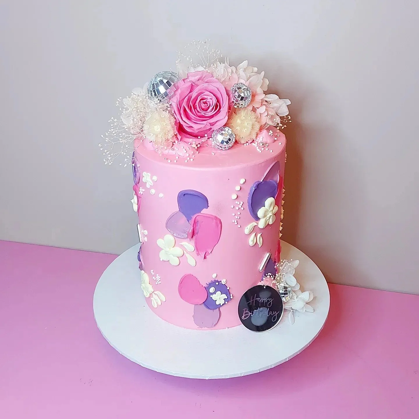 Pink birthday cake with white and purple decorations, topped with flowers, disco balls, and a 'Happy Birthday' topper on a white cake board against a pink and white background.