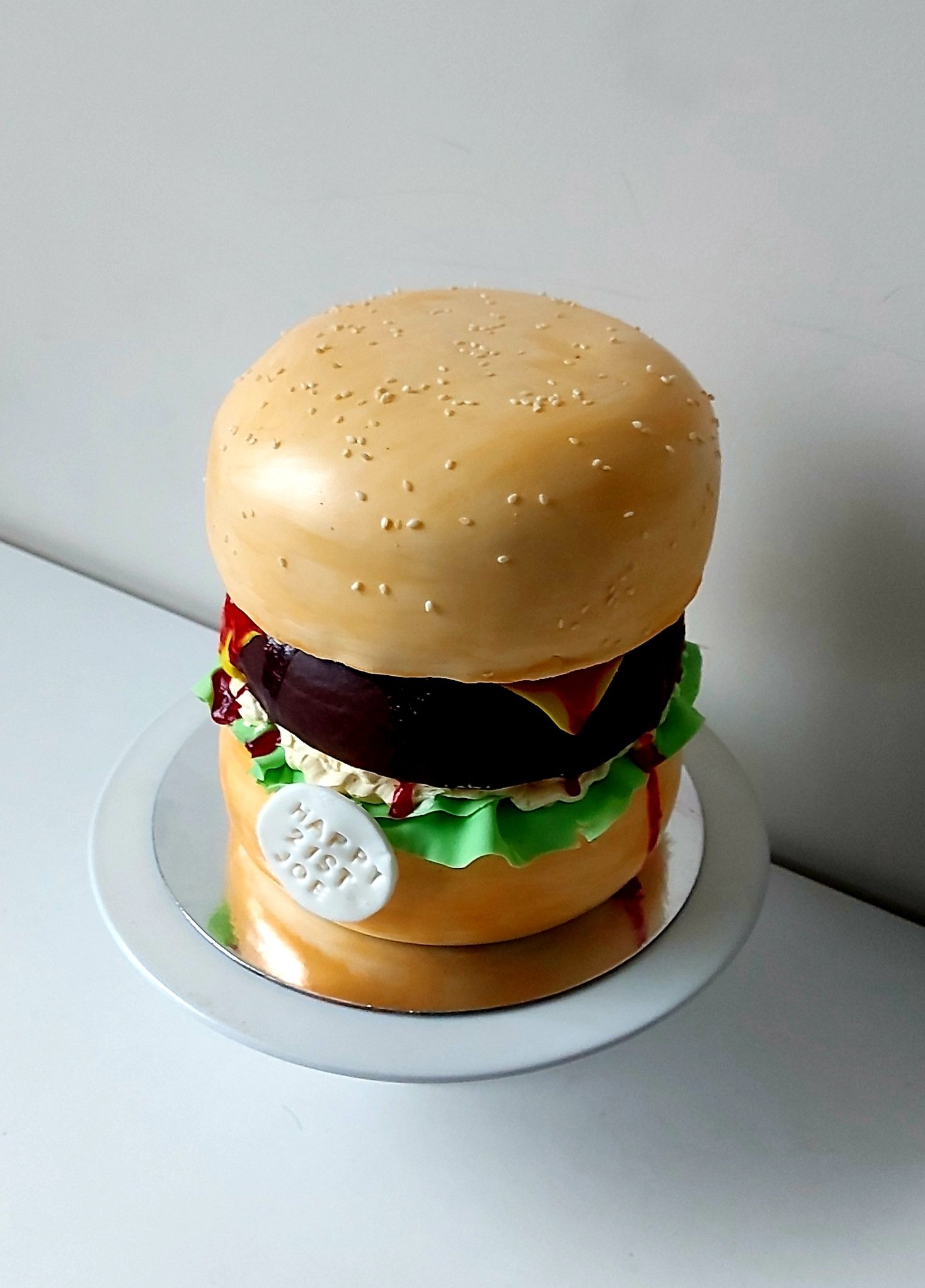 Cake shaped like a hamburger with detailed cake layers resembling a bun, lettuce, tomato, cheese, beef patty, and condiments, decorated with white edible pearls and a white edible badge with text.