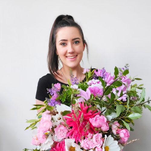 A young woman with dark brown hair in a high ponytail, smiling and holding a large bouquet of pink and purple flowers with green leaves.