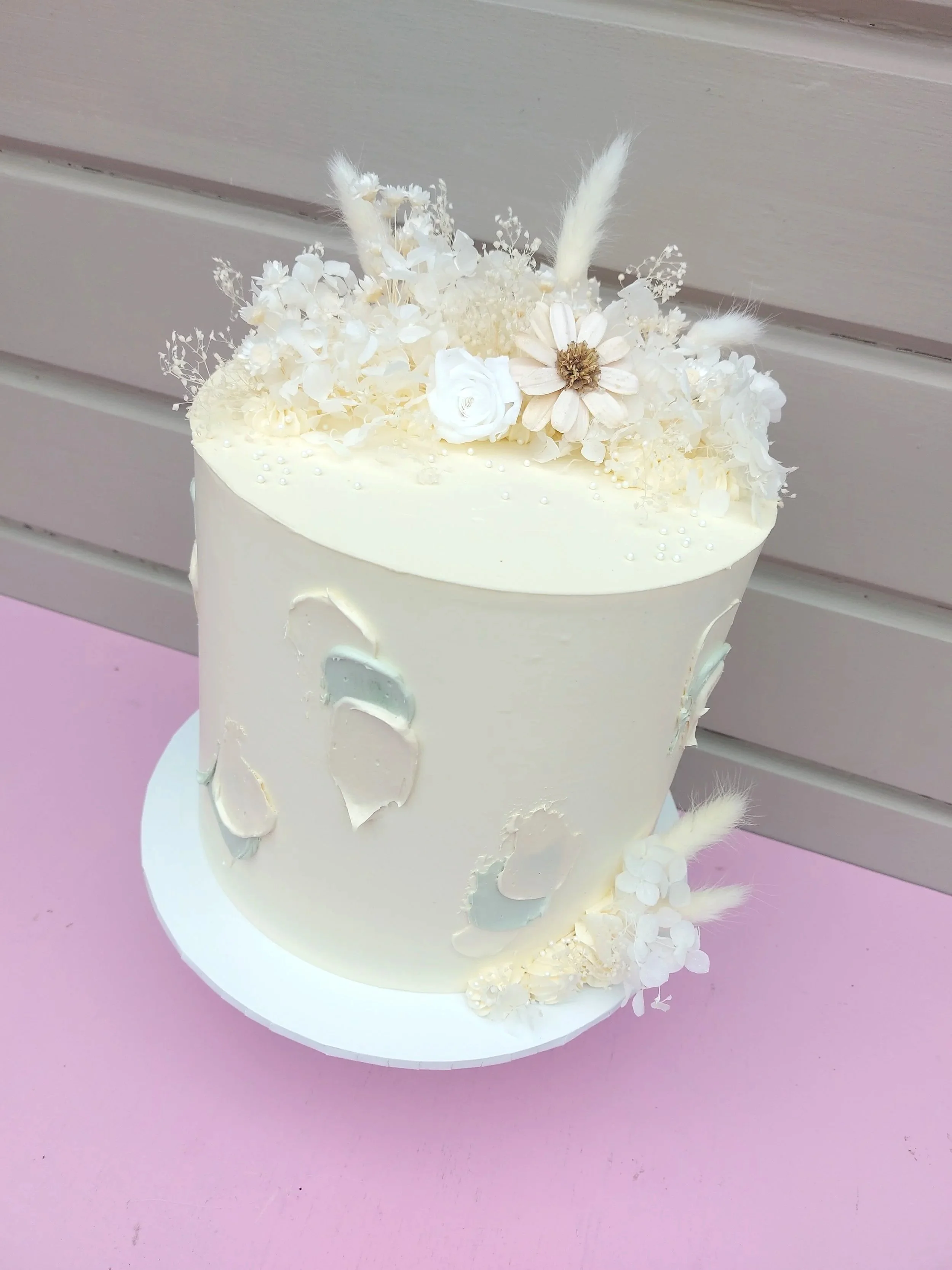 A white wedding cake decorated with white and cream-colored flowers and feathers, placed on a white cake stand with a pink surface underneath.