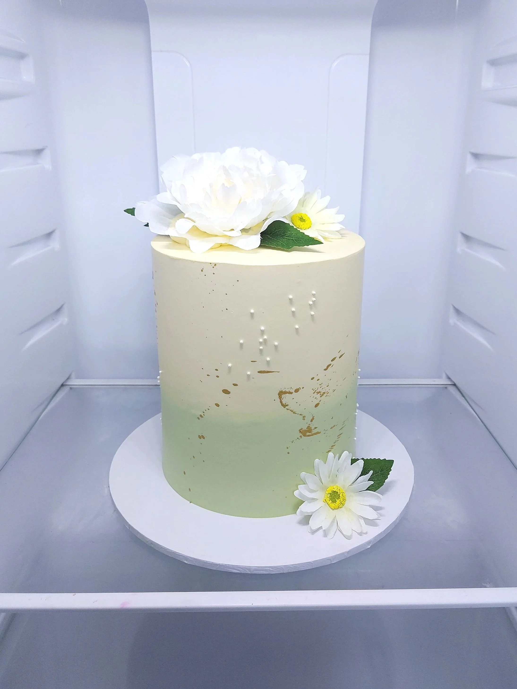 A white, round cake decorated with white flowers and green leaves, placed in a white display case with flowers on the inside.