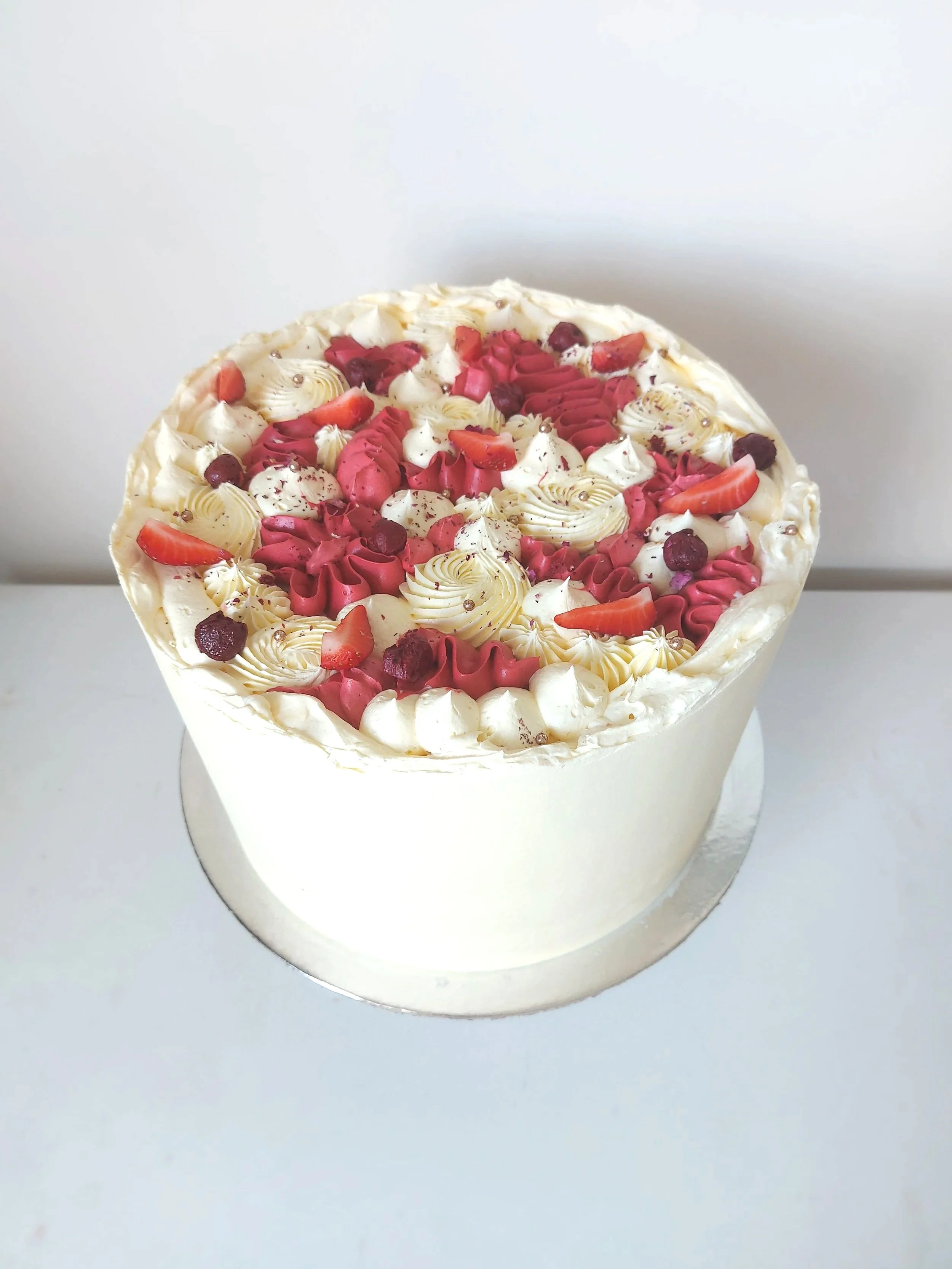 A round cake decorated with white, pink, and red icing and various berries, placed on a white surface with a plain light background.