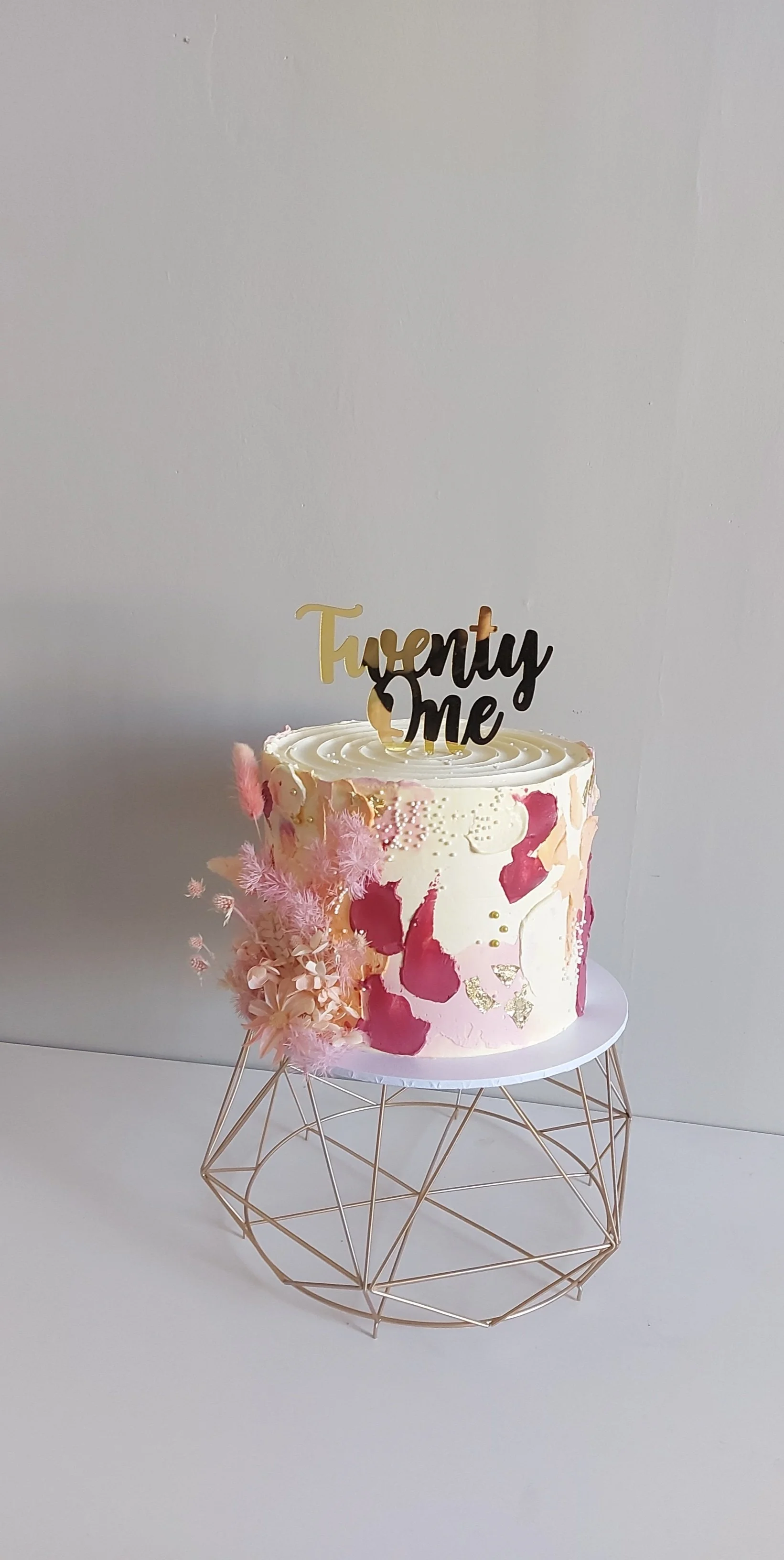 A decorative birthday cake with pink and gold accents, floral decorations on the side, and a 'Twenty One' topper on top, displayed on a wire stand against a plain white background.