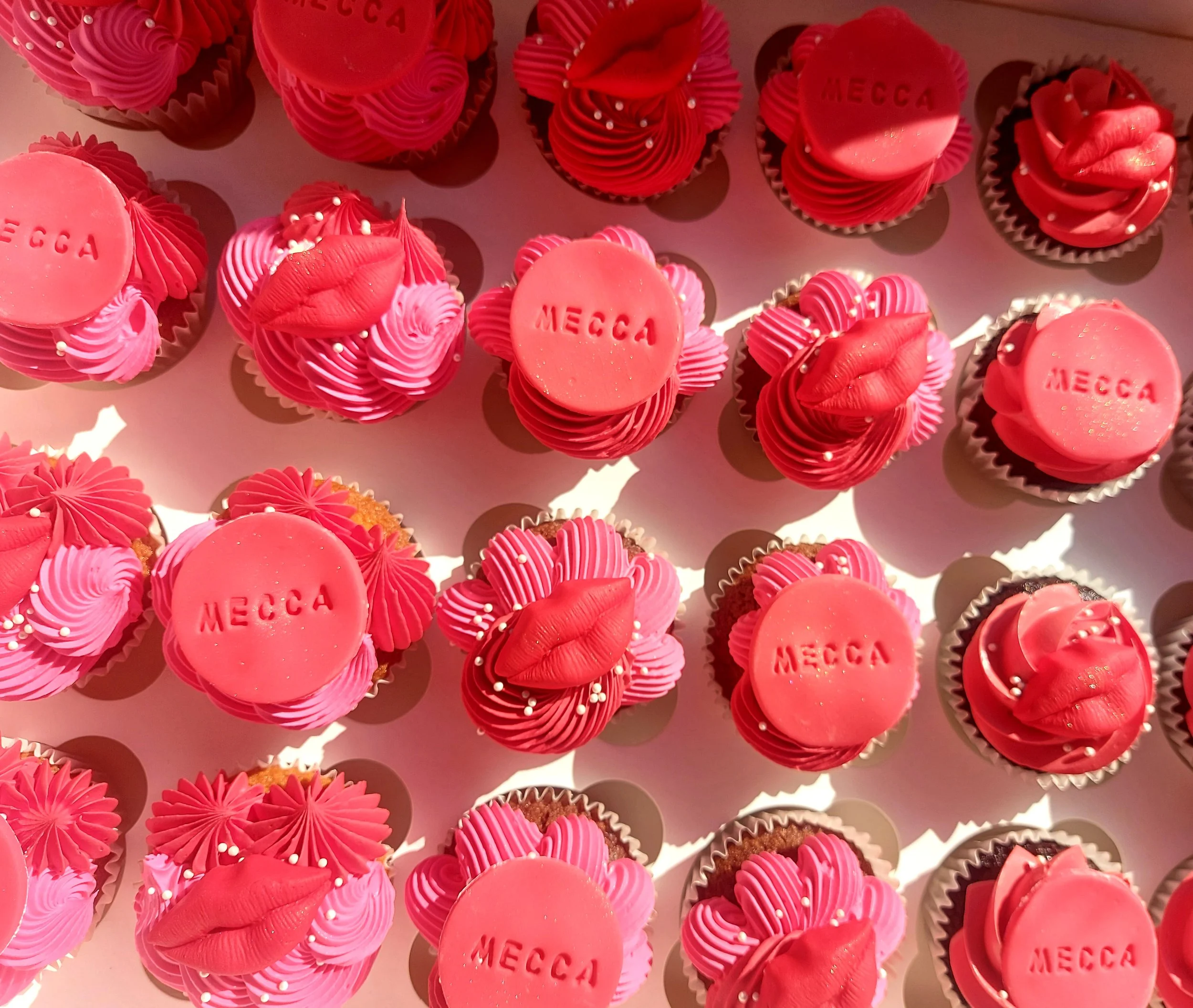 Multiple pink cupcakes decorated with pink frosting, some with white sprinkles, and topped with pink lips or round pink decorations with the word 'MECCA' engraved on them. The cupcakes are arranged on a white surface.