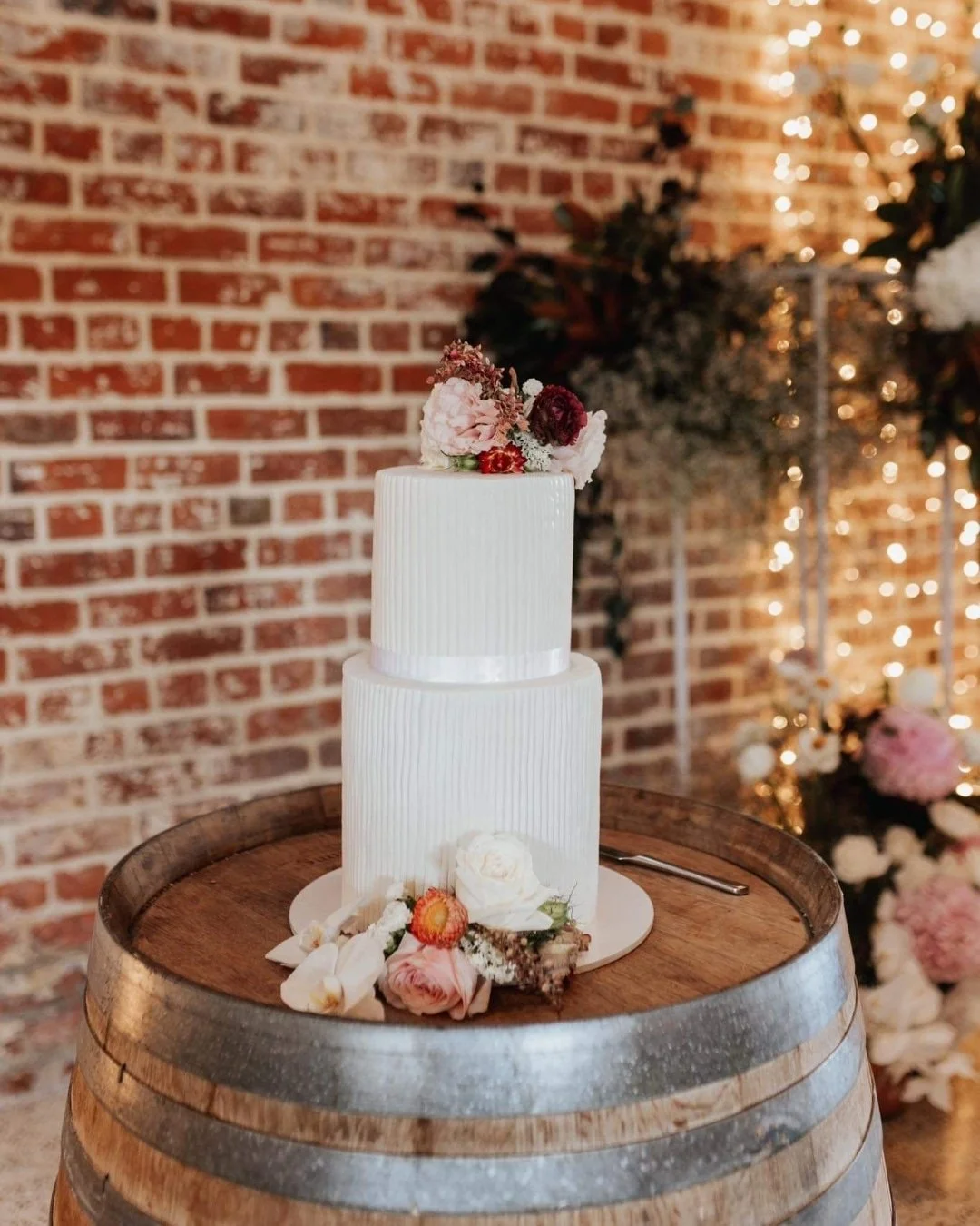 A two-tier white wedding cake with a floral topper, placed on a wooden barrel, decorated with flowers at the base, against a brick wall with string lights and greenery in the background.