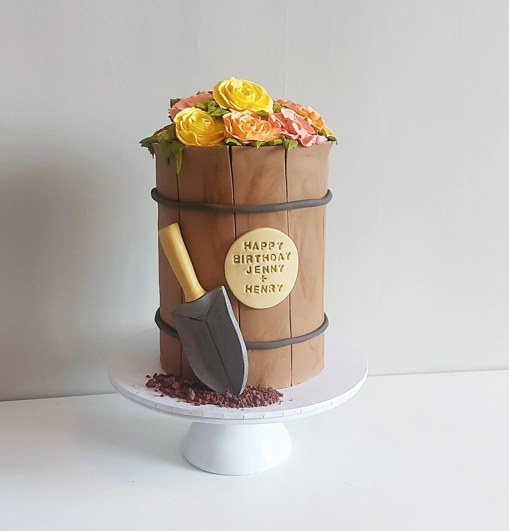 Cake shaped like a flower bucket with yellow and pink roses on top, a black shovel cake decoration, and a white chocolate plaque with a birthday message for Jenny and Henry, on a white cake stand.