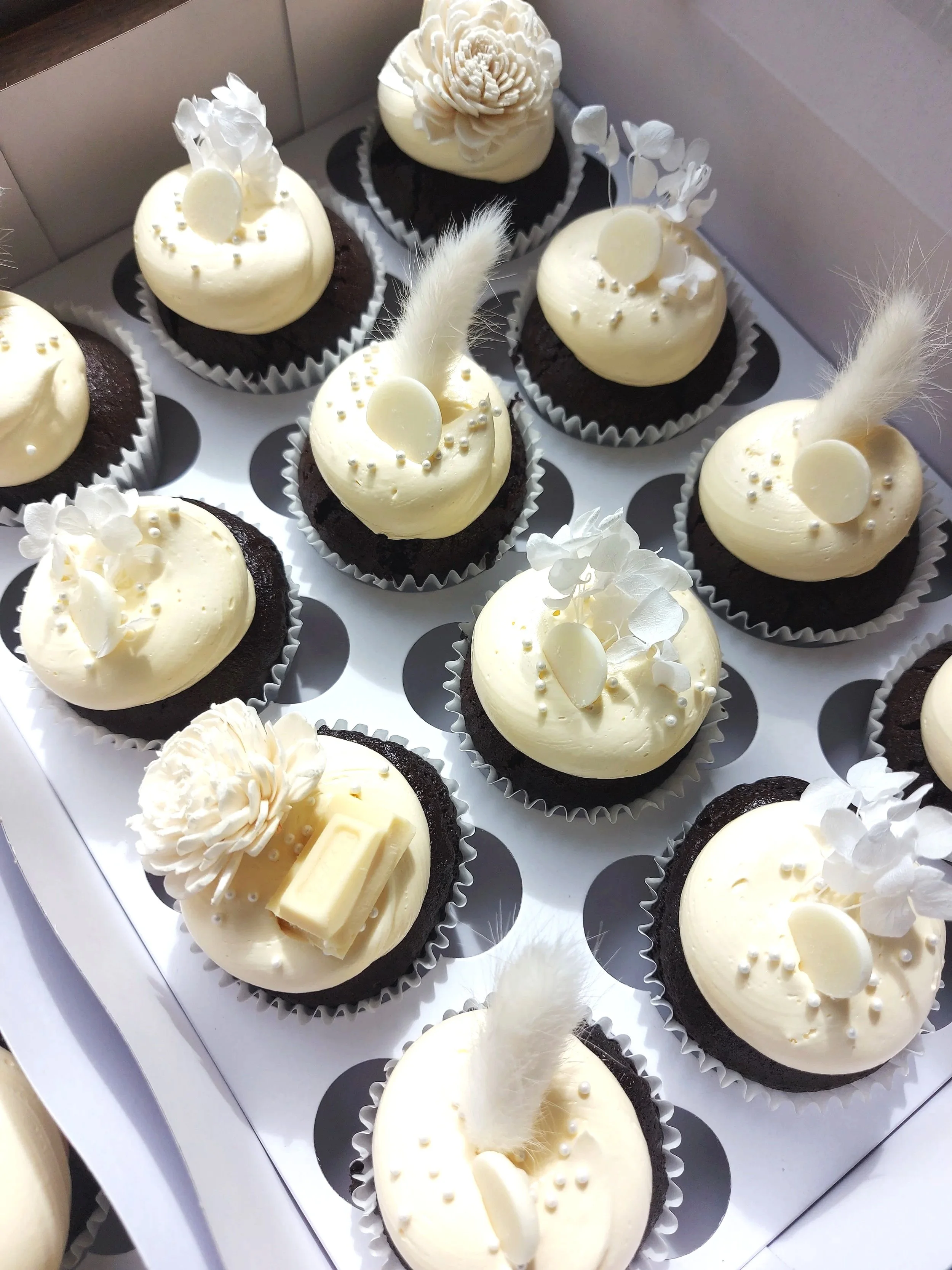 A box of decorated chocolate cupcakes with white frosting, decorative white flowers, feathers, and white chocolate pieces.