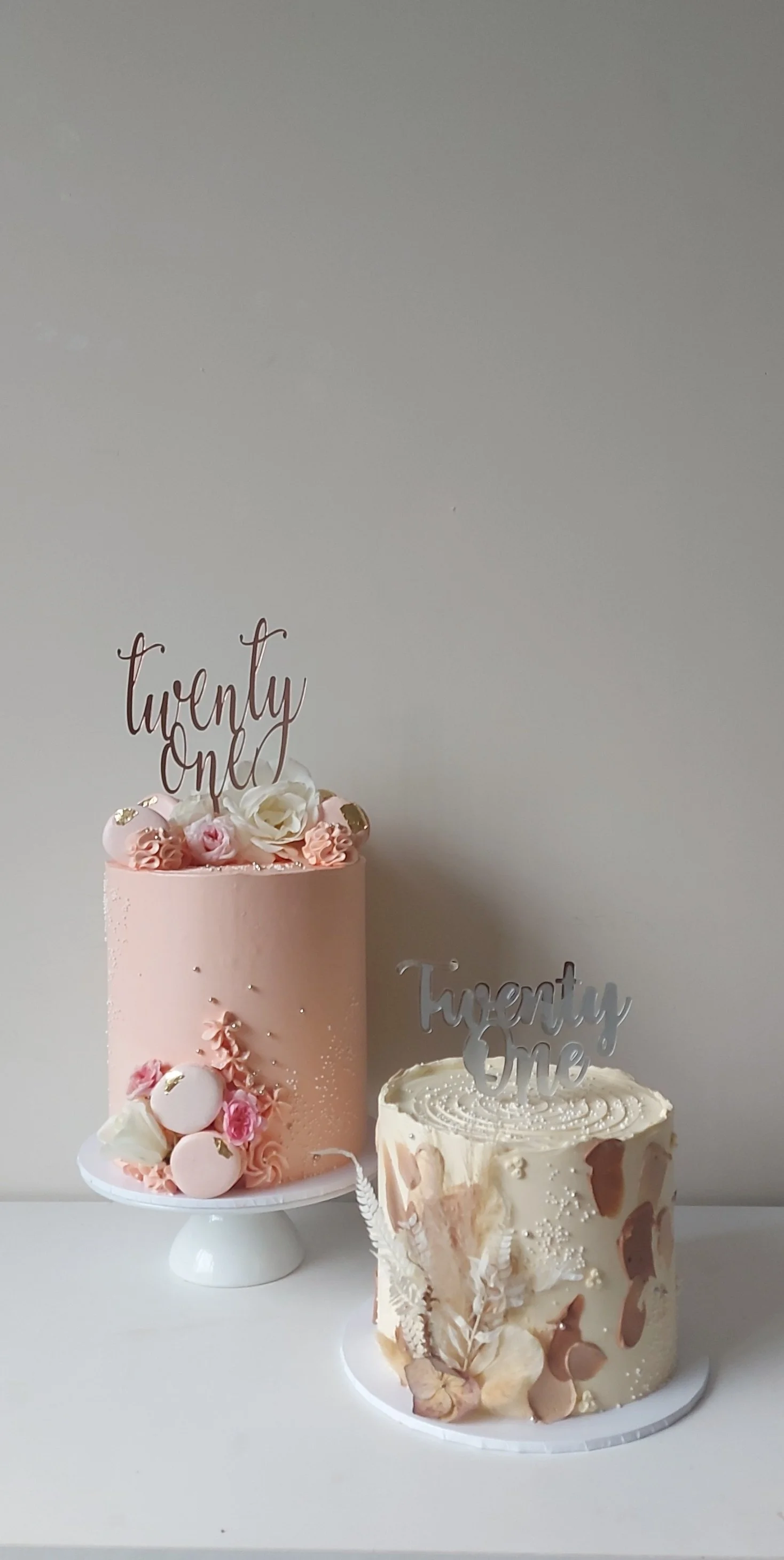 Two decorated celebration cakes with floral and pearl embellishments, one pink with a 'twenty one' topper, and another white with decorative accents, set against a plain gray wall.