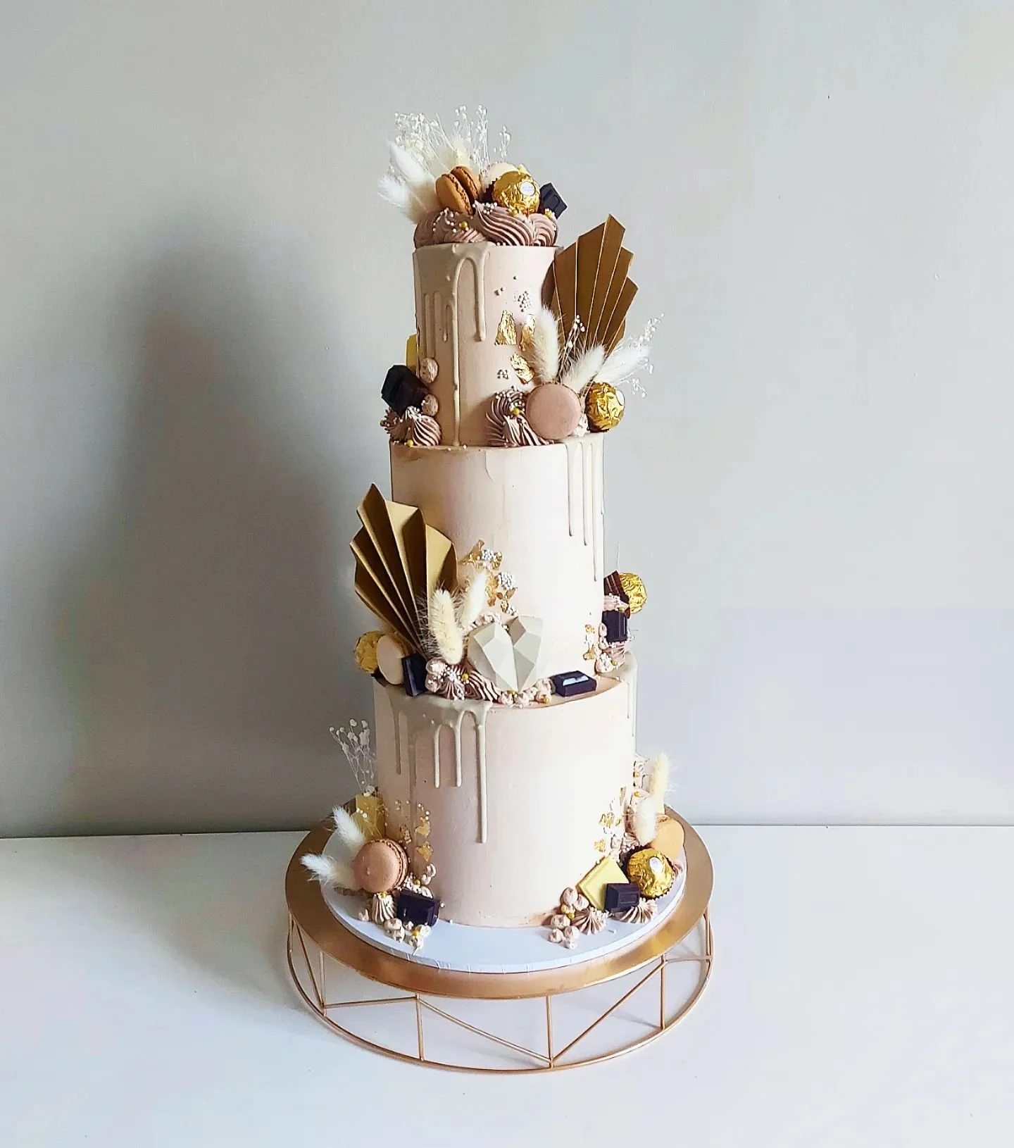 Three-tiered wedding cake decorated with gold, white, and pink accents, featuring chocolates, macarons, feathered decorations, and geometric embellishments, placed on a copper-colored wire stand against a plain background.