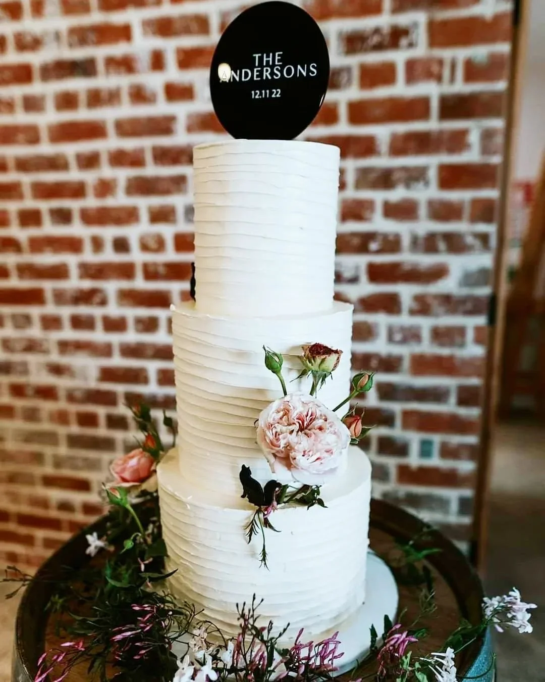 3-tier white wedding cake decorated with pink and white flowers and greenery, topped with a black circular topper with white text that reads 'The Andersons' and a date, set against a brick wall background.