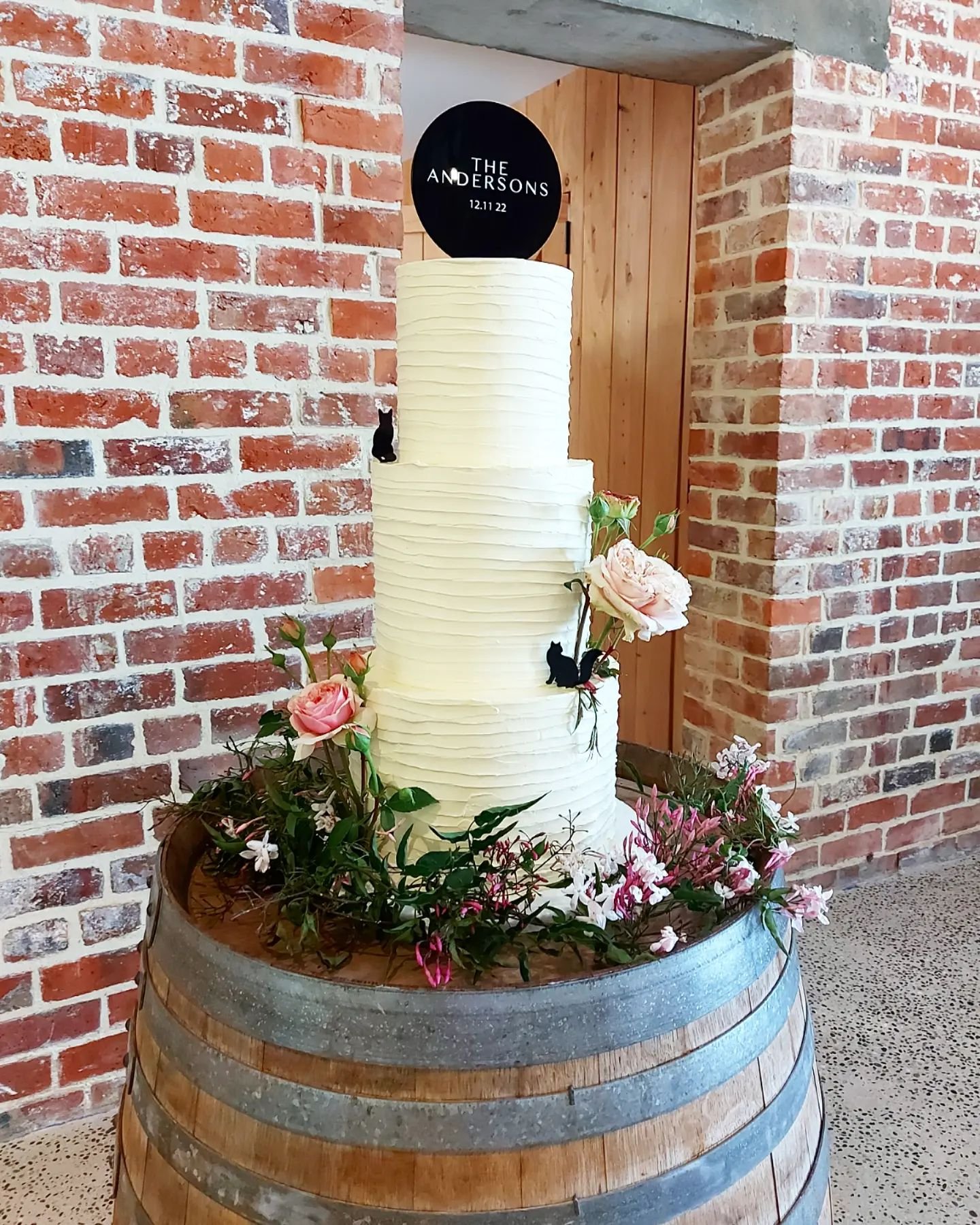 A tall white wedding cake with minimal decoration, surrounded by pink and white flowers, placed inside a wooden barrel against a brick wall background.