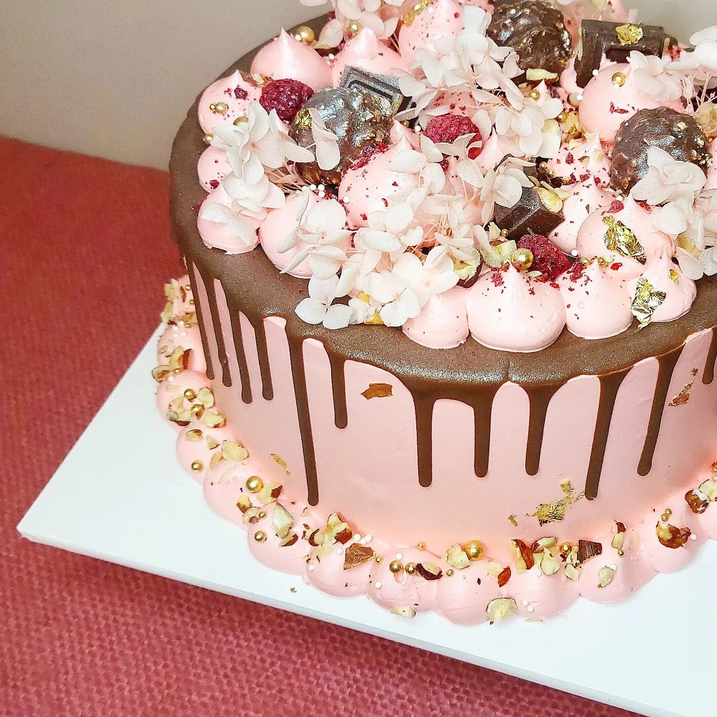 A pink birthday cake with chocolate drizzle, topped with white and pink frosting, dried raspberries, edible gold leaf, and white flower decorations.
