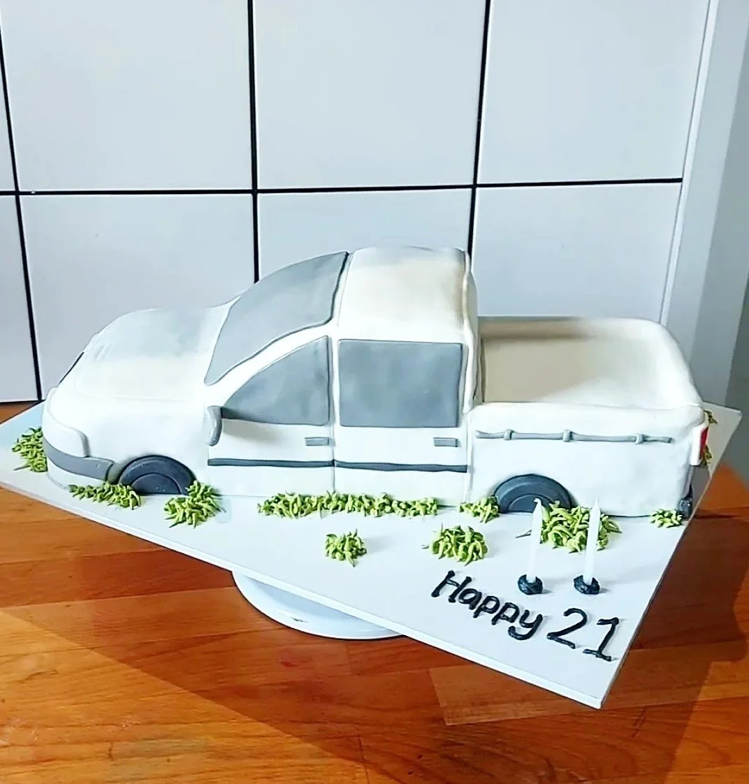 A cake decorated to look like a white pickup truck, with green icing bushes around it, on a white cake board. The cake has a note saying "Happy 21".