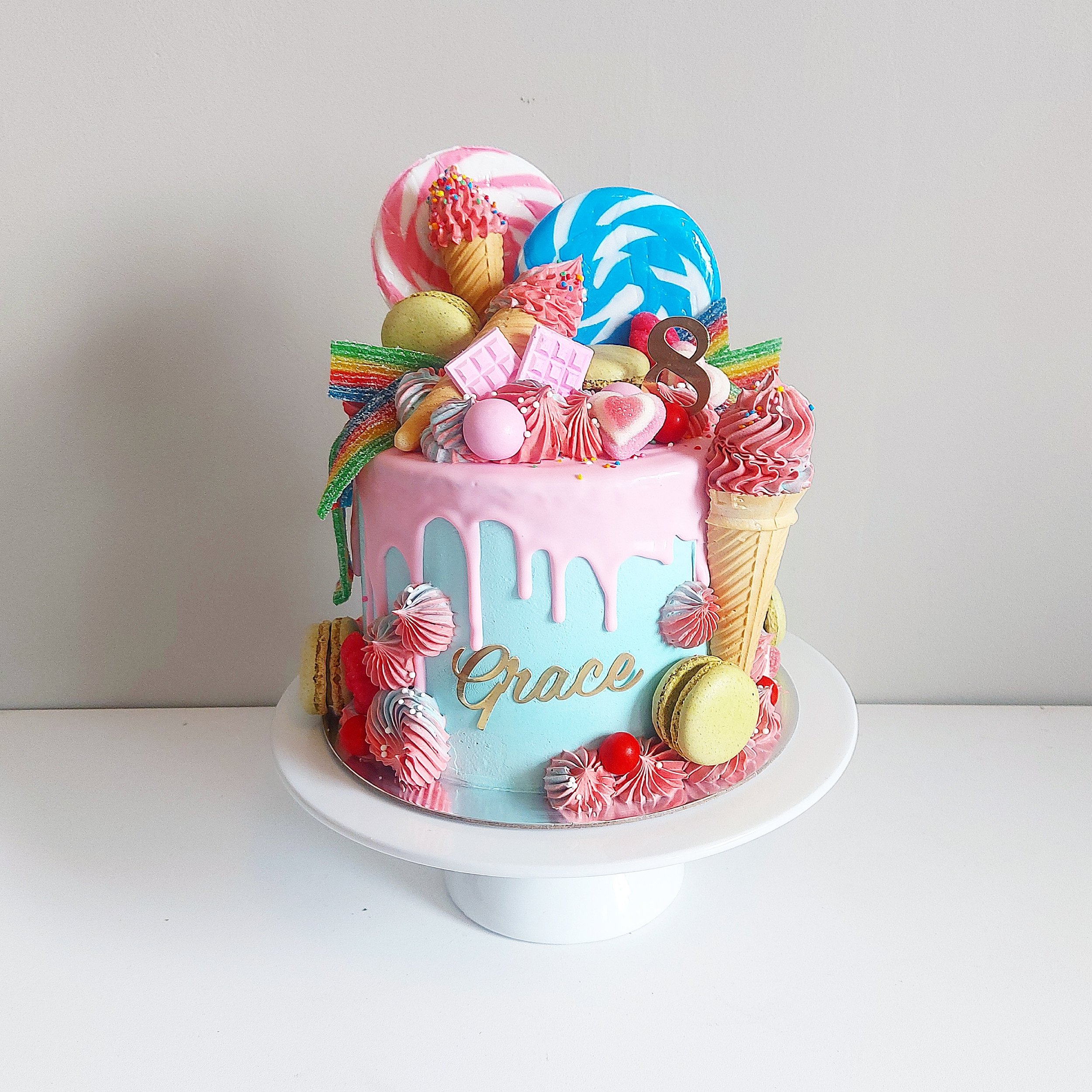 Colorful birthday cake decorated with candies, macarons, pink and blue frosting, rainbow strips, and an ice cream cone with pink frosting and sprinkles.