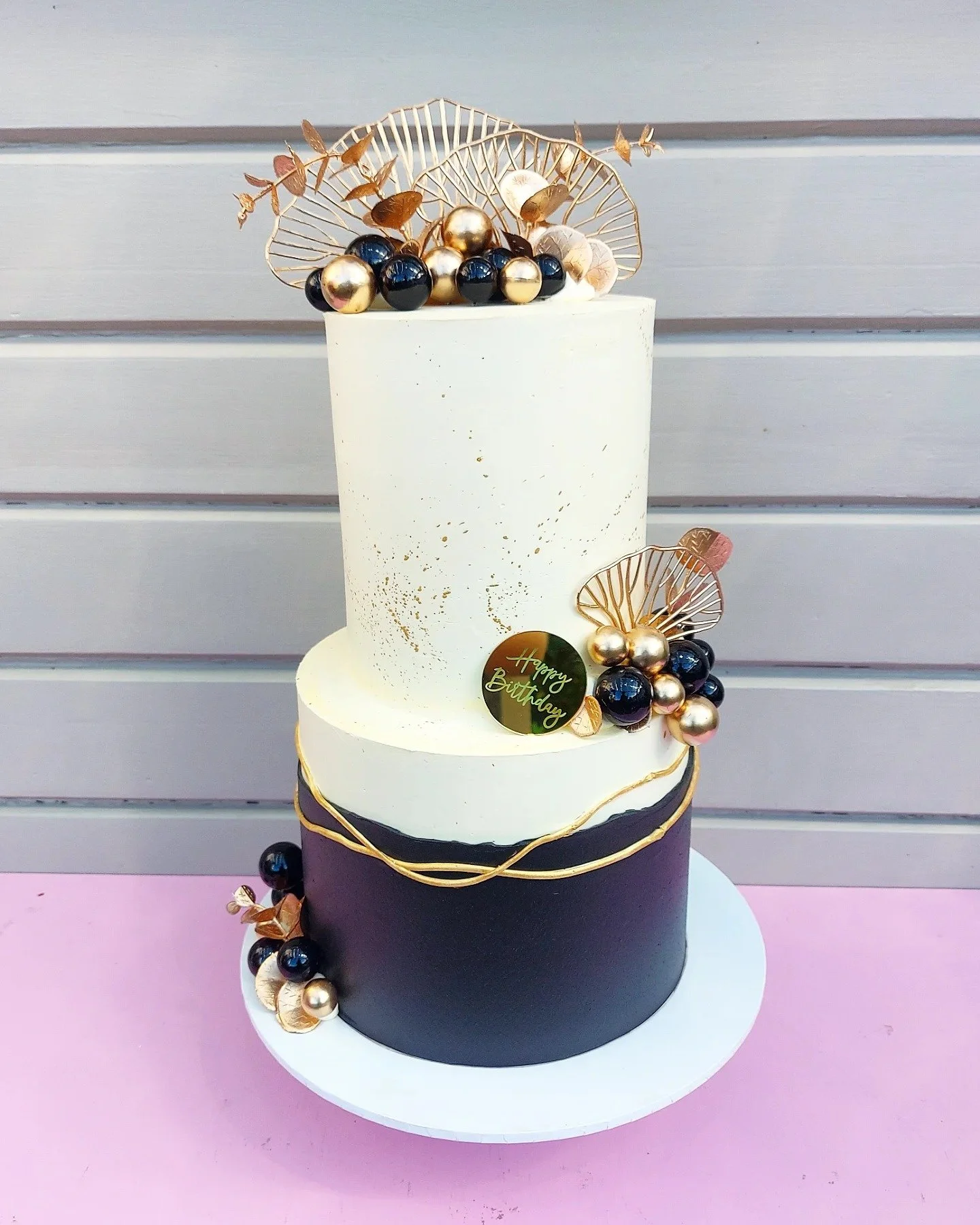 A three-tier birthday cake with black, white, and gold decorations, topped with gold, black, and white beads, gold leaves, and a small black gift tag that reads "Happy Birthday" against a gray wooden wall background.