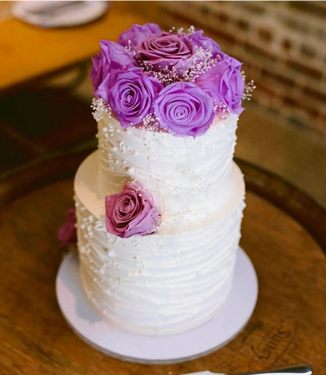 A three-tier white wedding cake decorated with purple roses and small white decorative elements, placed on a wooden surface.