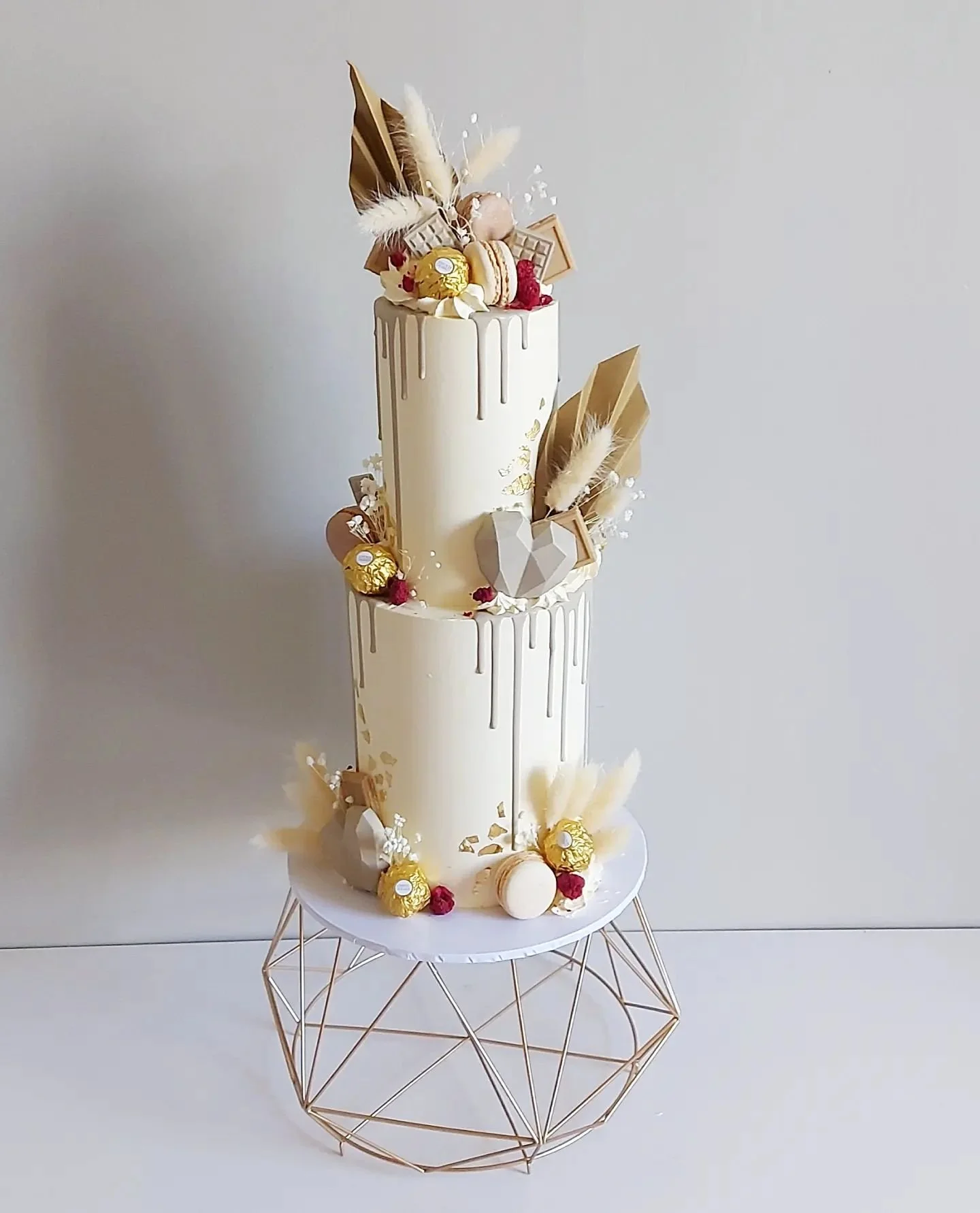 Elegant three-tier white cake decorated with chocolates, macarons, red berries, and feathers, placed on a modern geometric metal stand against a plain background.