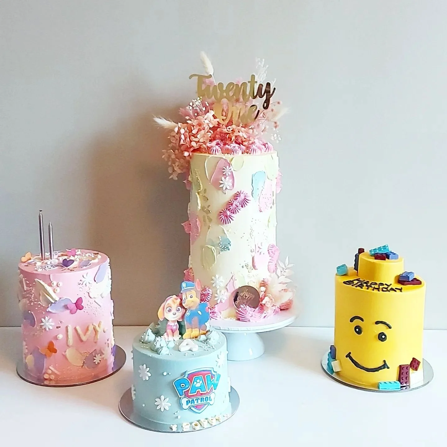 A collection of colorful birthday cakes with various themes and decorations, including a pink cake with hearts and butterfly decorations, a gray "Paw Patrol" themed cake, a large white cake with pink flowers and butterflies, and a yellow lego-themed 
