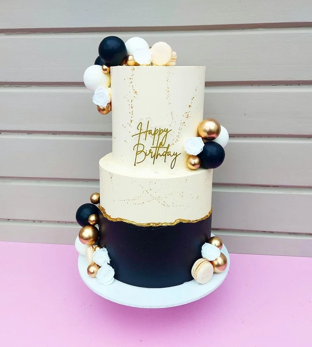 Two-tiered birthday cake with black, white, and gold decorations and a "Happy Birthday" message in gold lettering, topped with spherical accents and small floral decorations.
