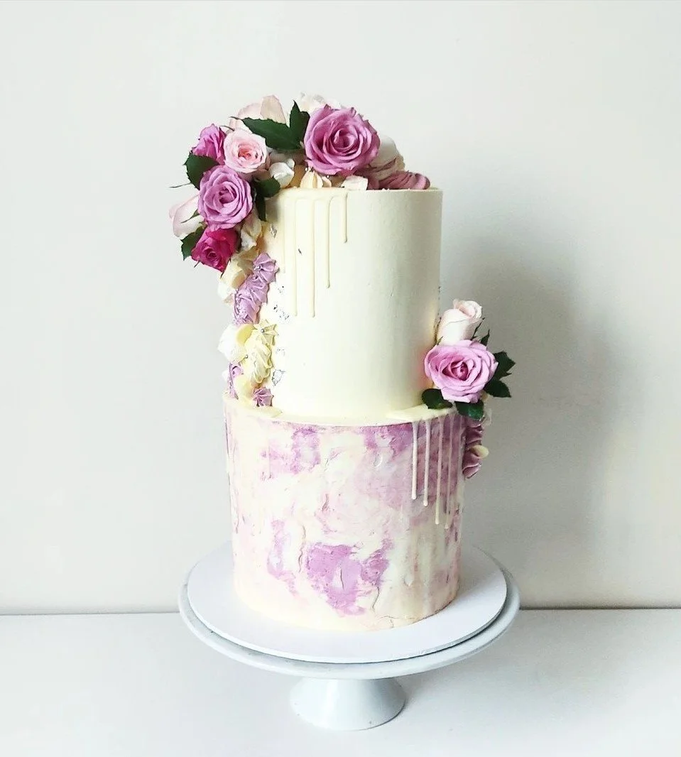 Two-tiered cake with pink and white marbled icing, decorated with pink and purple roses and white flowers, with a white drip design on the top tier, displayed on a white cake stand against a plain background.