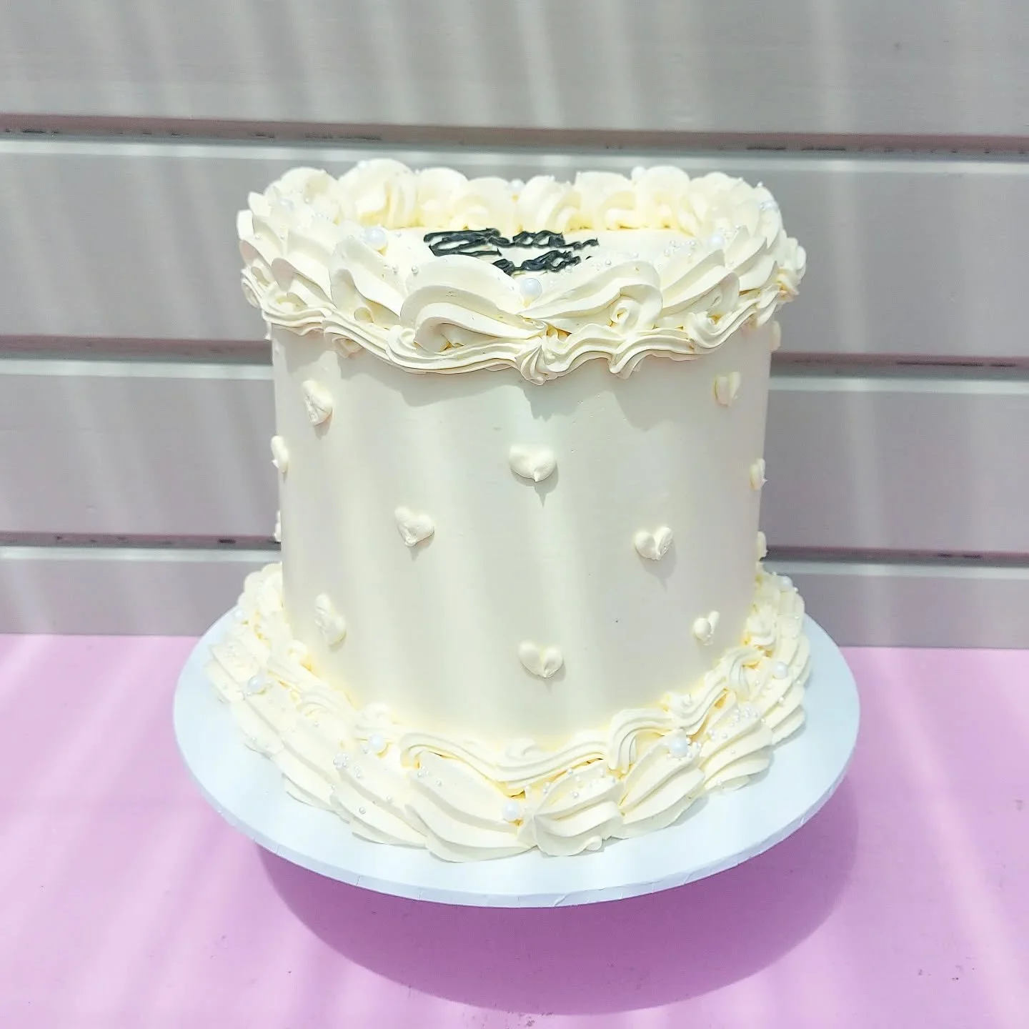 A white cake decorated with white icing swirls, small white heart-shaped candies, and silver edible beads. The top layer has black writing, and both layers are surrounded by decorative icing on the base.