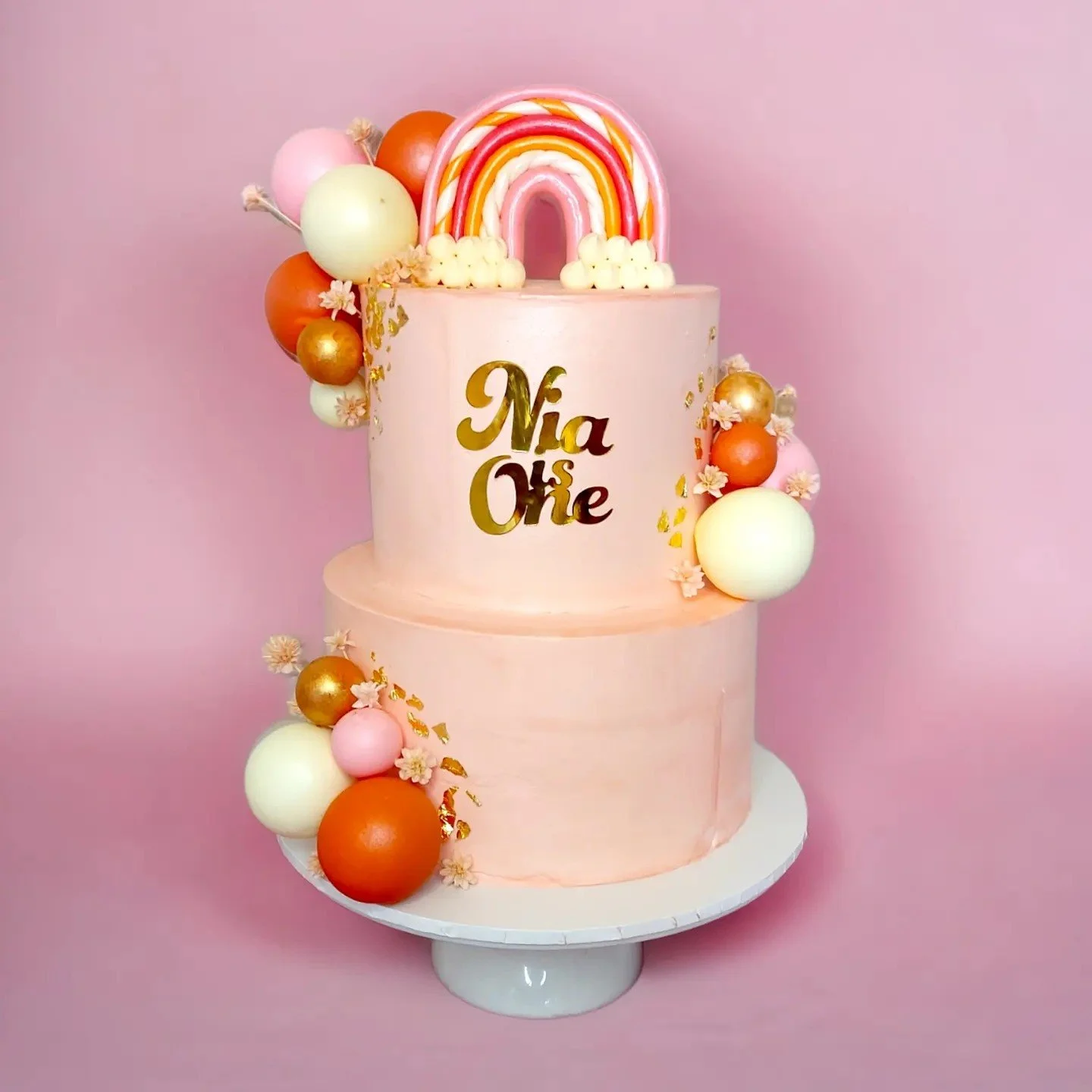 Pink two-tiered birthday cake with gold lettering 'Nia 1st', decorated with pastel-colored spheres, small flowers, and a rainbow topper, on a white cake stand against a pink background.